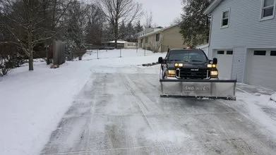 Black vehicle with a snowplow attached, clearing snow from a driveway in a snowy residential area.