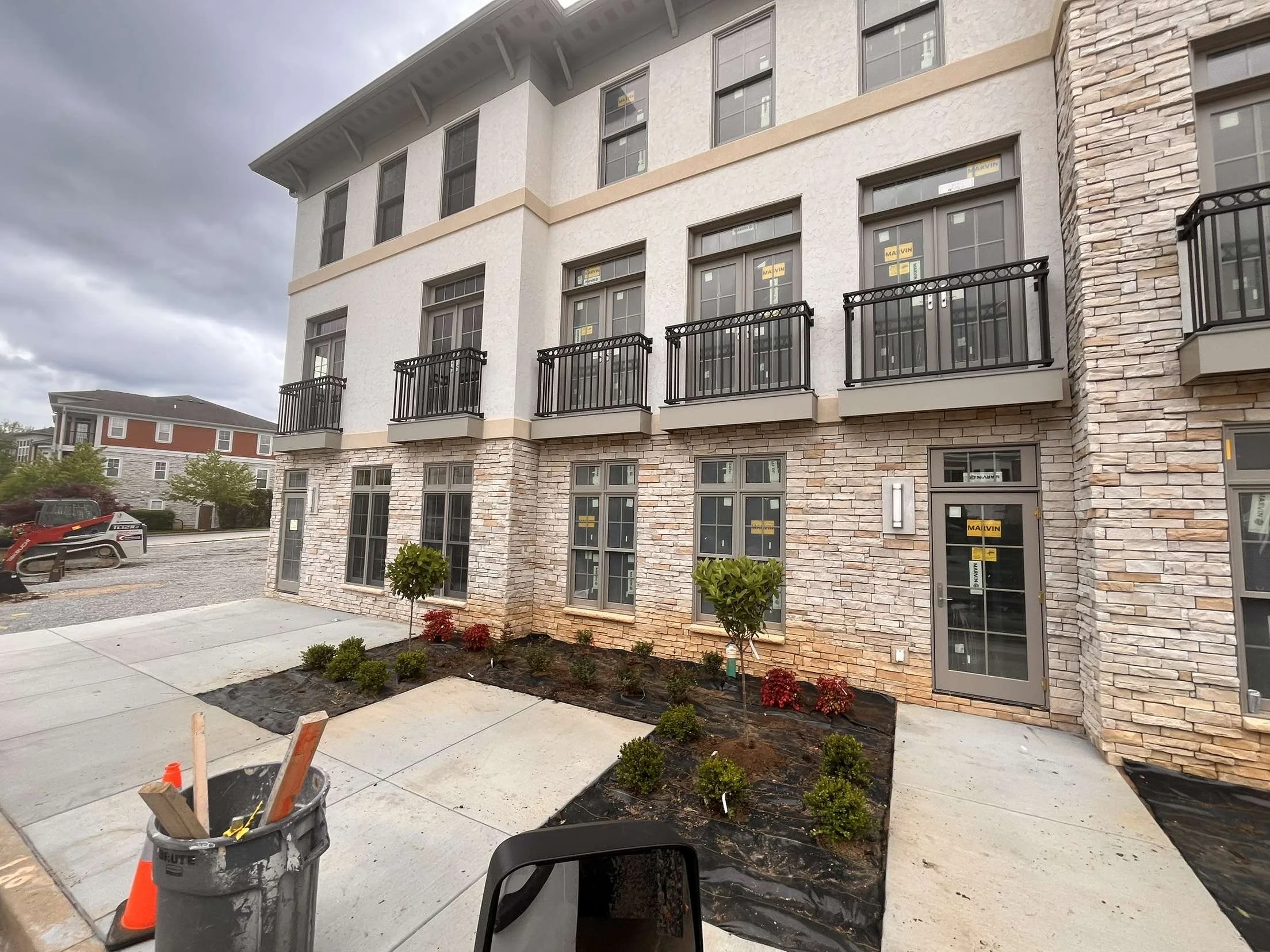 Newly constructed multi-story building with stone and white stucco exterior, black metal balconies, and window coverings labeled 'MA	extsubscript{V}IN'; landscaped area in front with small trees and bushes; cloudy sky overhead.
