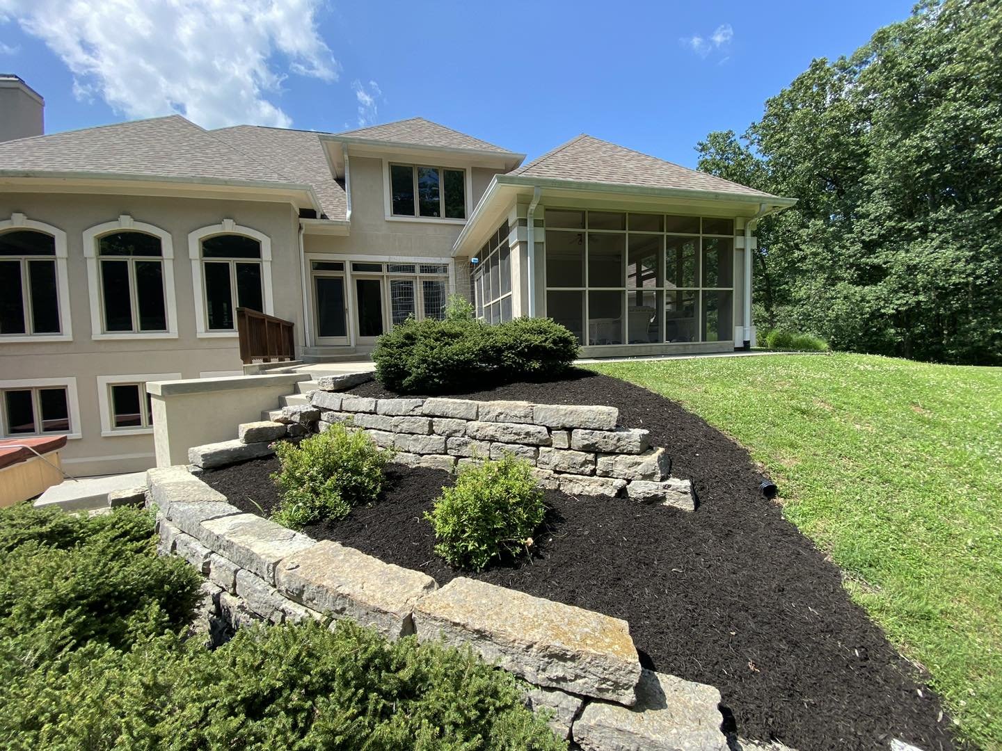Backyard with landscaped garden, stone retaining wall, green shrubs, and a screened porch attached to a two-story house with beige exterior and multiple windows, under a partly cloudy sky.