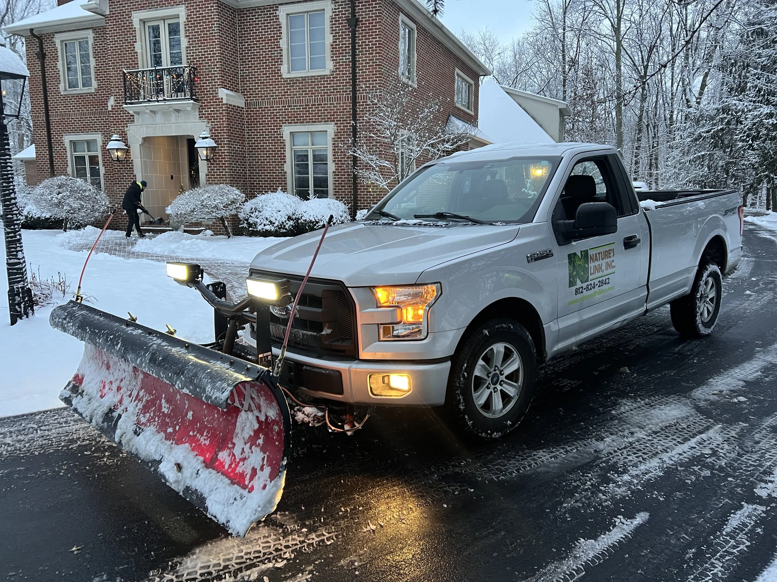 A pickup truck equipped with a snow plow clearing snow from a driveway in front of a brick house during winter.