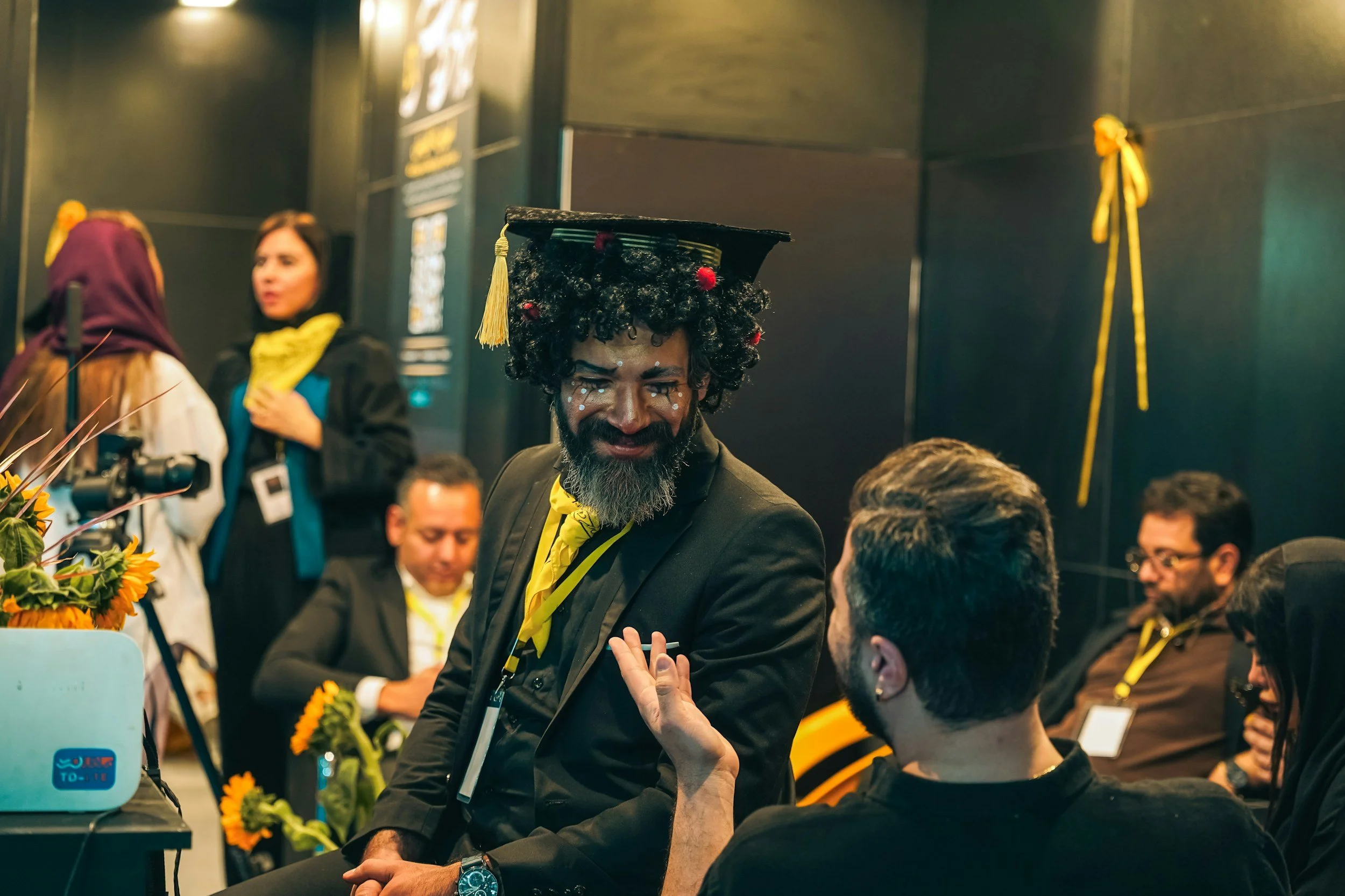 A man with curly black hair, painted face, and a black academic cap with yellow tassel is smiling and talking to another man in a formal setting. The man with the face paint is dressed in a black suit, with yellow accents, and is seated among others who are also dressed formally. There are people in the background, some standing and some sitting, with yellow decorations and sunflowers visible.