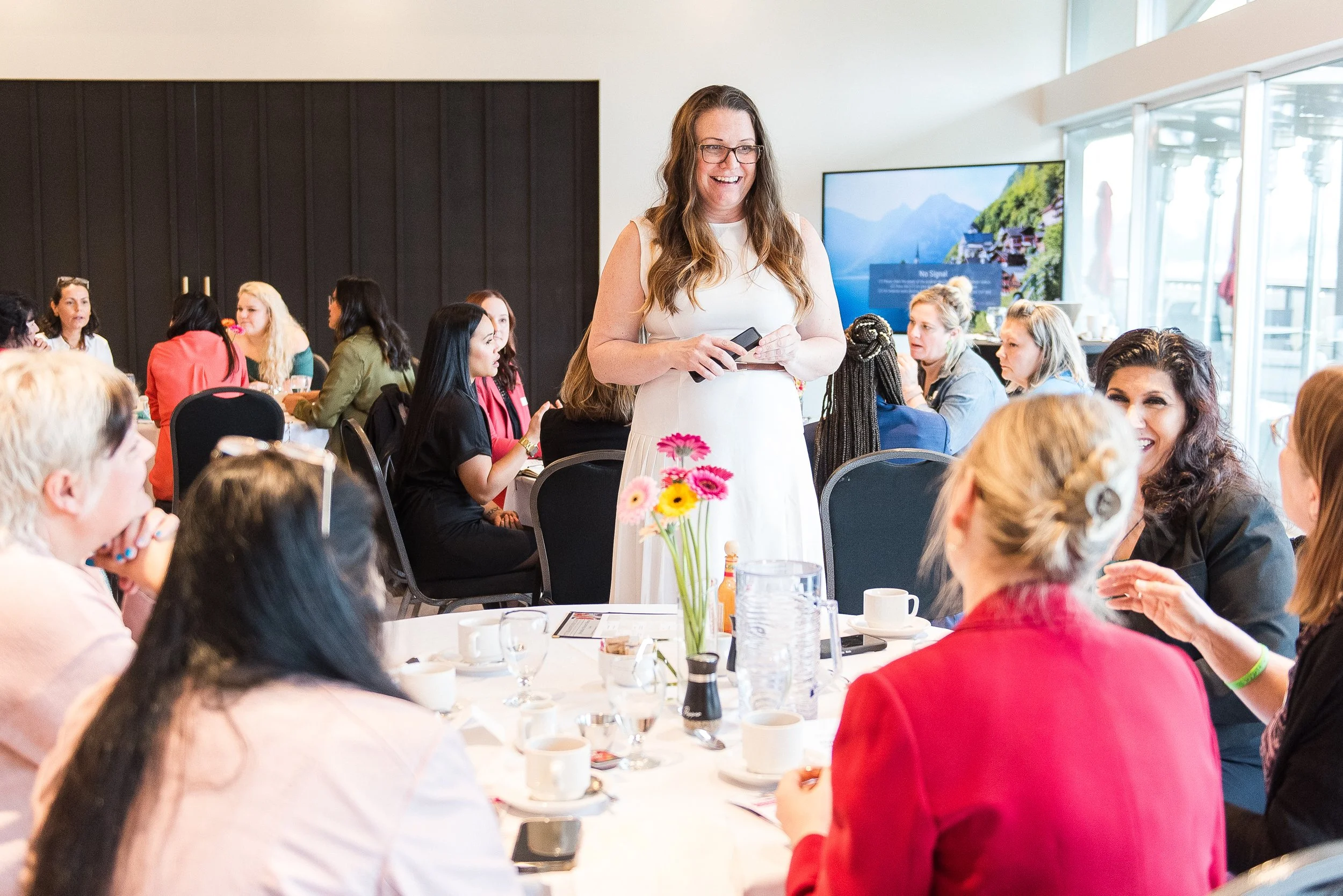 Meghan Simington in a white dress and glasses, holding a phone, standing at a round table during a conference or meeting, surrounded by other women sitting and engaging in conversation at a Female Forward Collective Breakfast