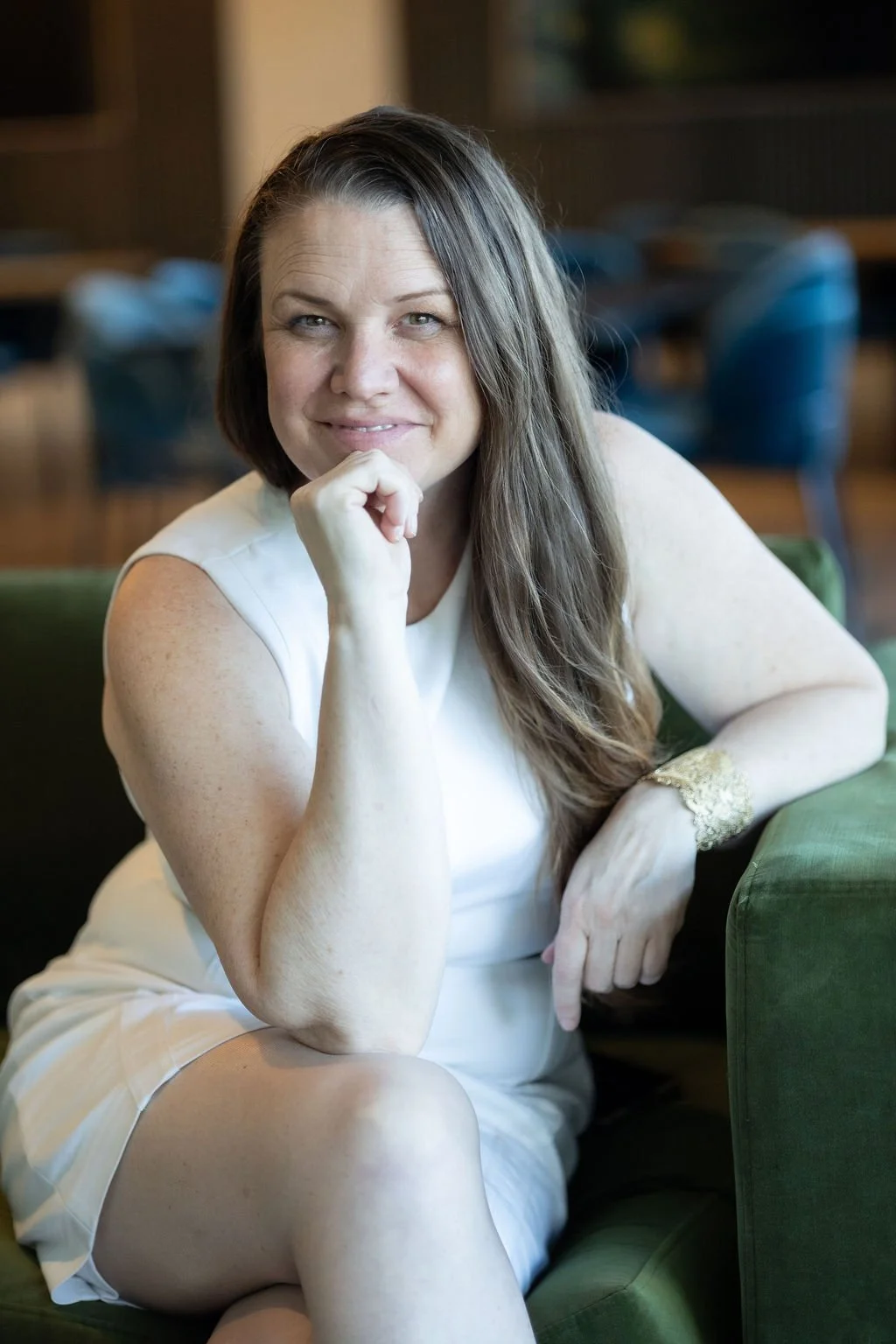 Meghan Simington smiling with her chin resting on her hand, sitting on a green couch in a room with wooden furniture.