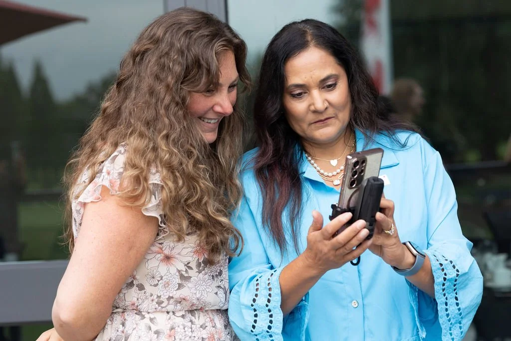 Two women looking at a smartphone together outdoors, one with curly hair and the other with straight black hair, wearing a blue blouse.