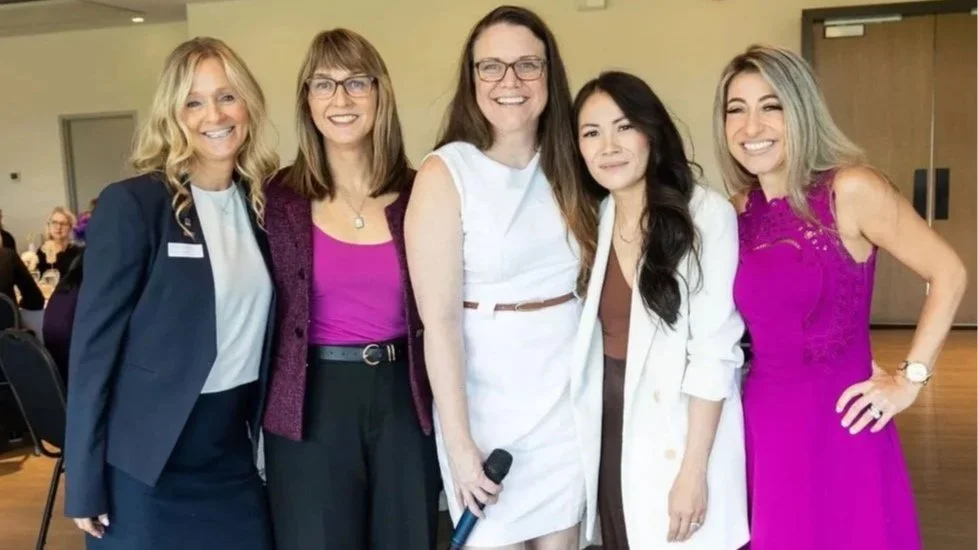 Five women smiling and posing together indoors at a social event, some holding a microphone.