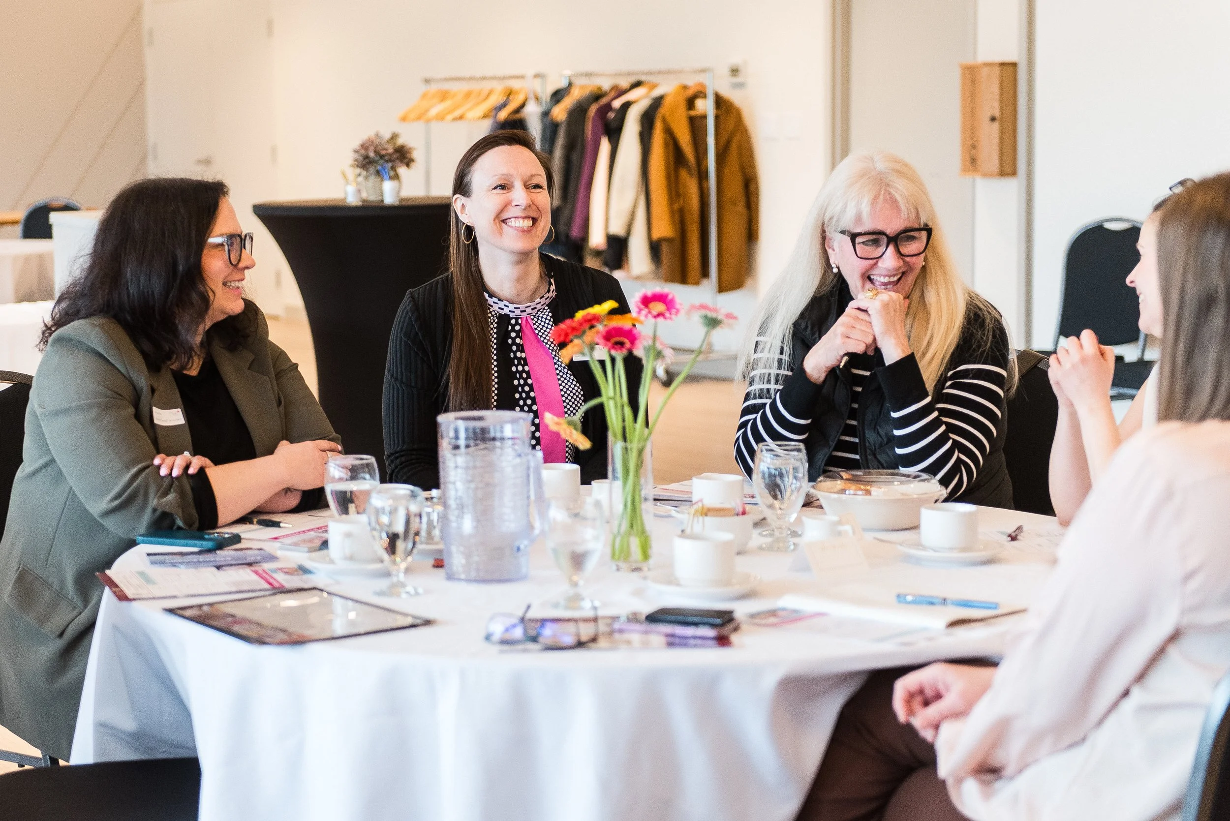 A group of five women sitting around a table in a meeting or conference room, smiling and engaging in conversation, with a vase of colorful flowers in the center.
