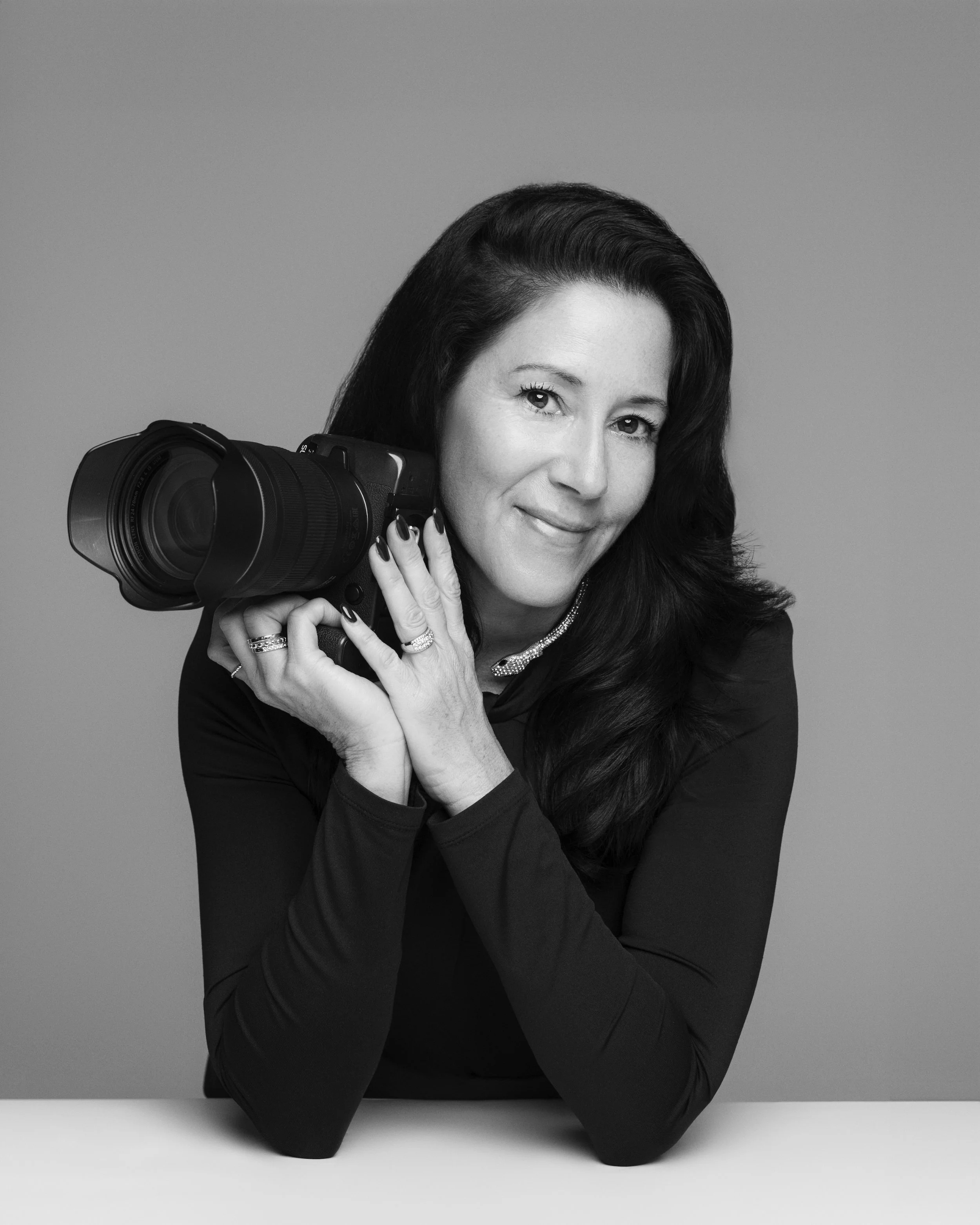 A woman with long dark hair holding a professional camera on her shoulder, smiling at the camera in a studio setting.