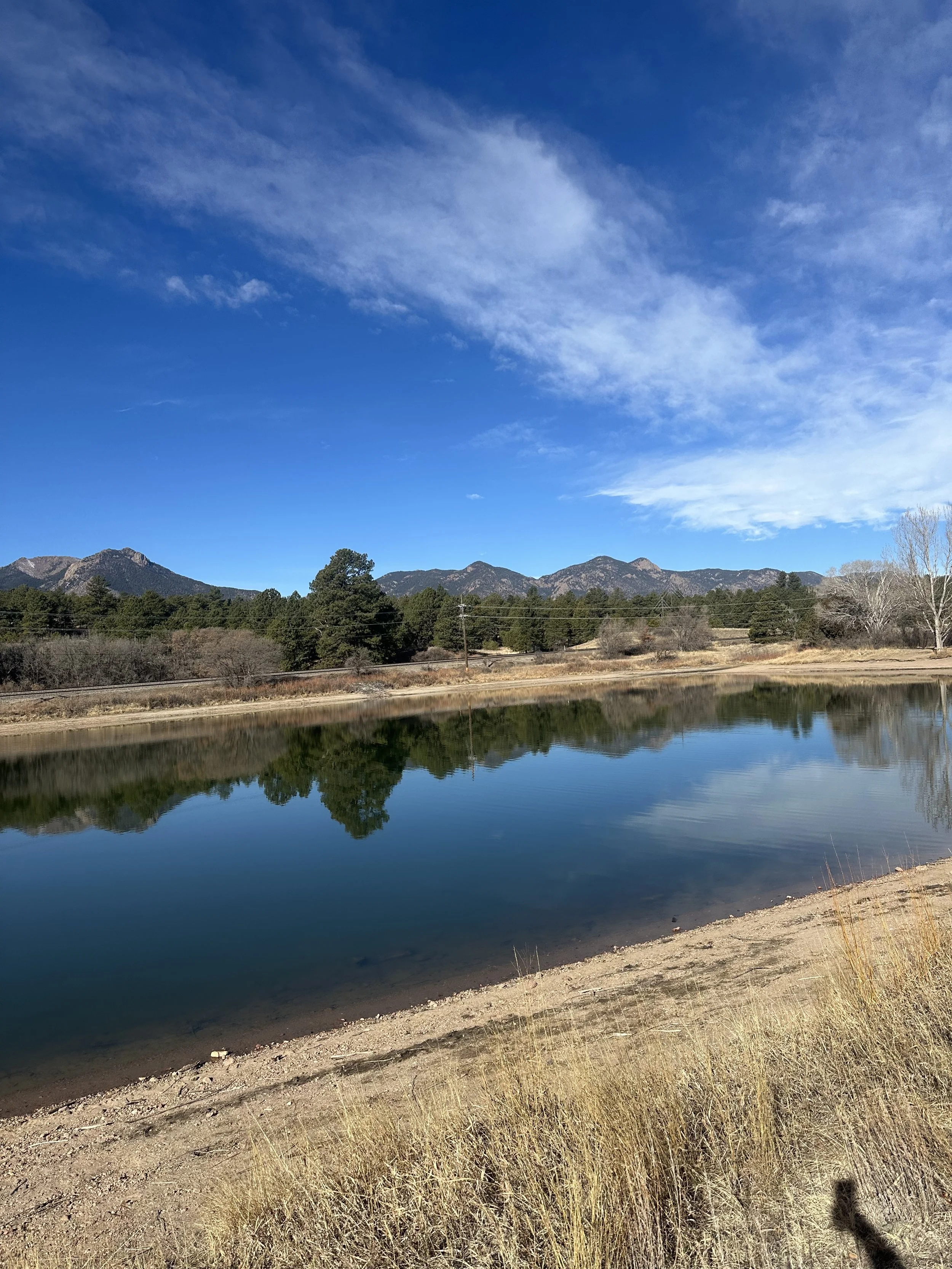 Scenic landscape featuring a calm lake, trees, mountains in the background, and a blue sky with scattered clouds.