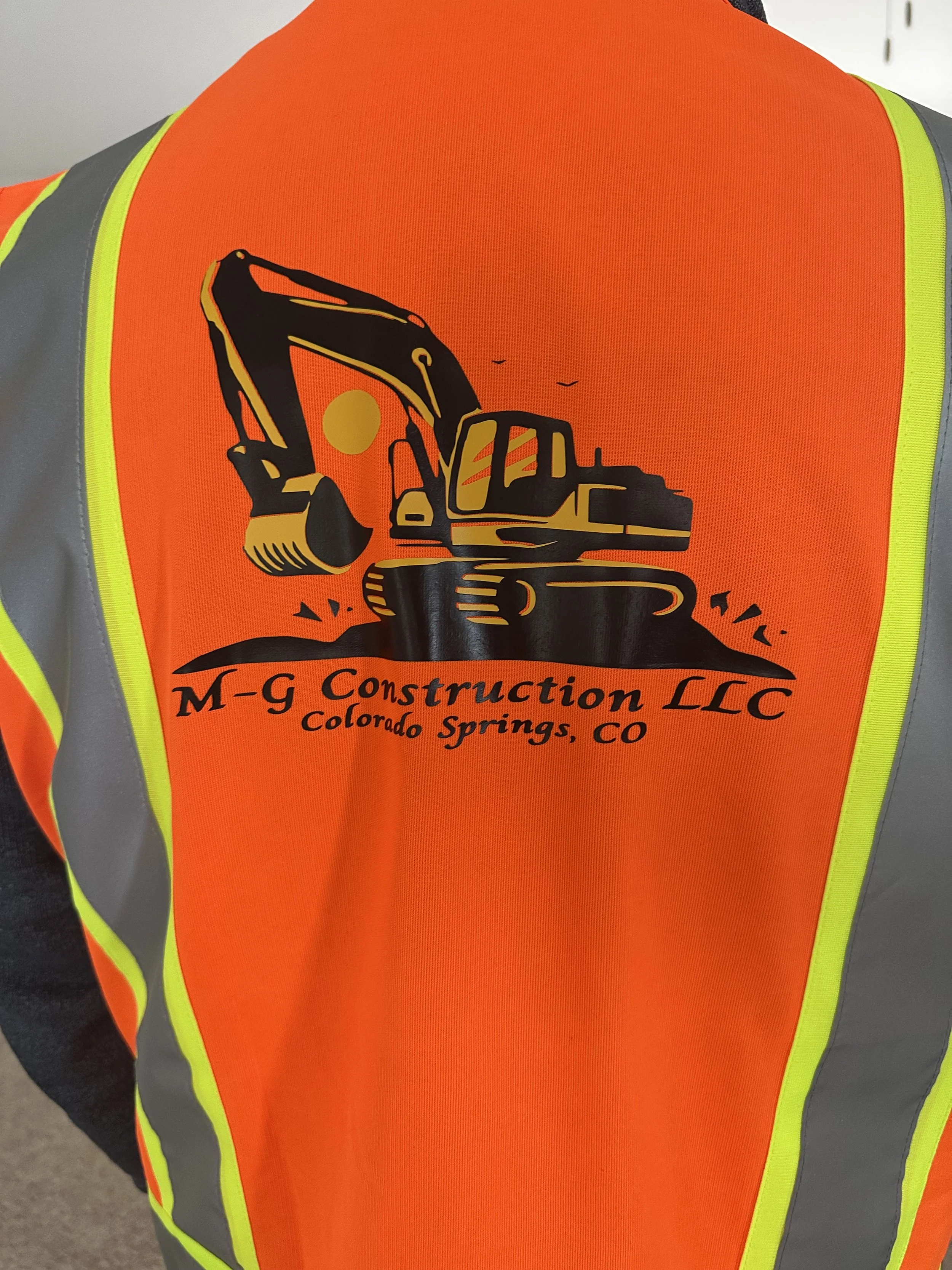 Back of a construction worker wearing a bright orange safety vest with reflective yellow and gray stripes. The vest features a logo for M-g Construction LLC, Colorado Springs, CO, with a black graphic of an excavator and the sun.