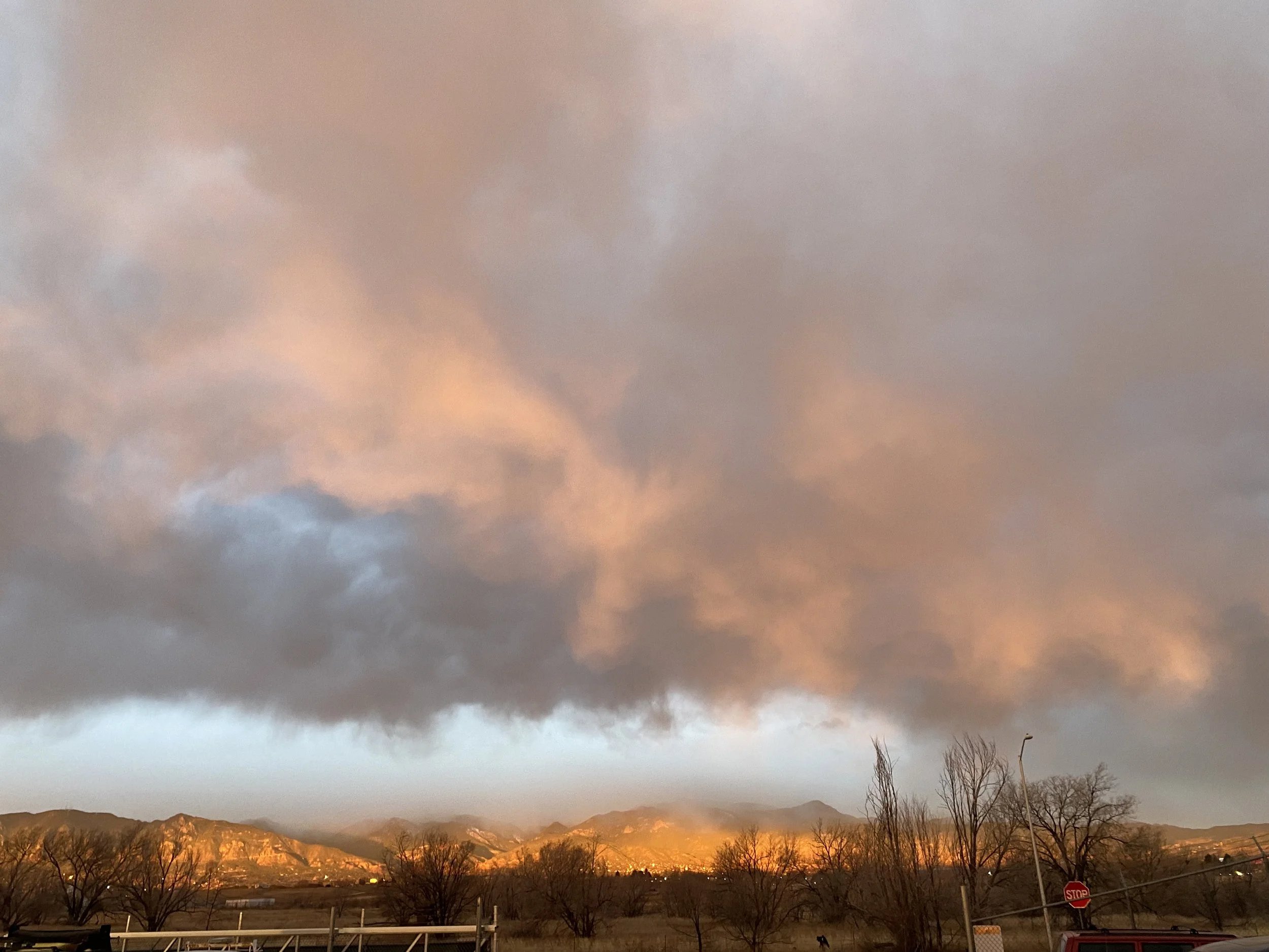 Sunset sky with pink and orange clouds over mountains and barren trees in the foreground.