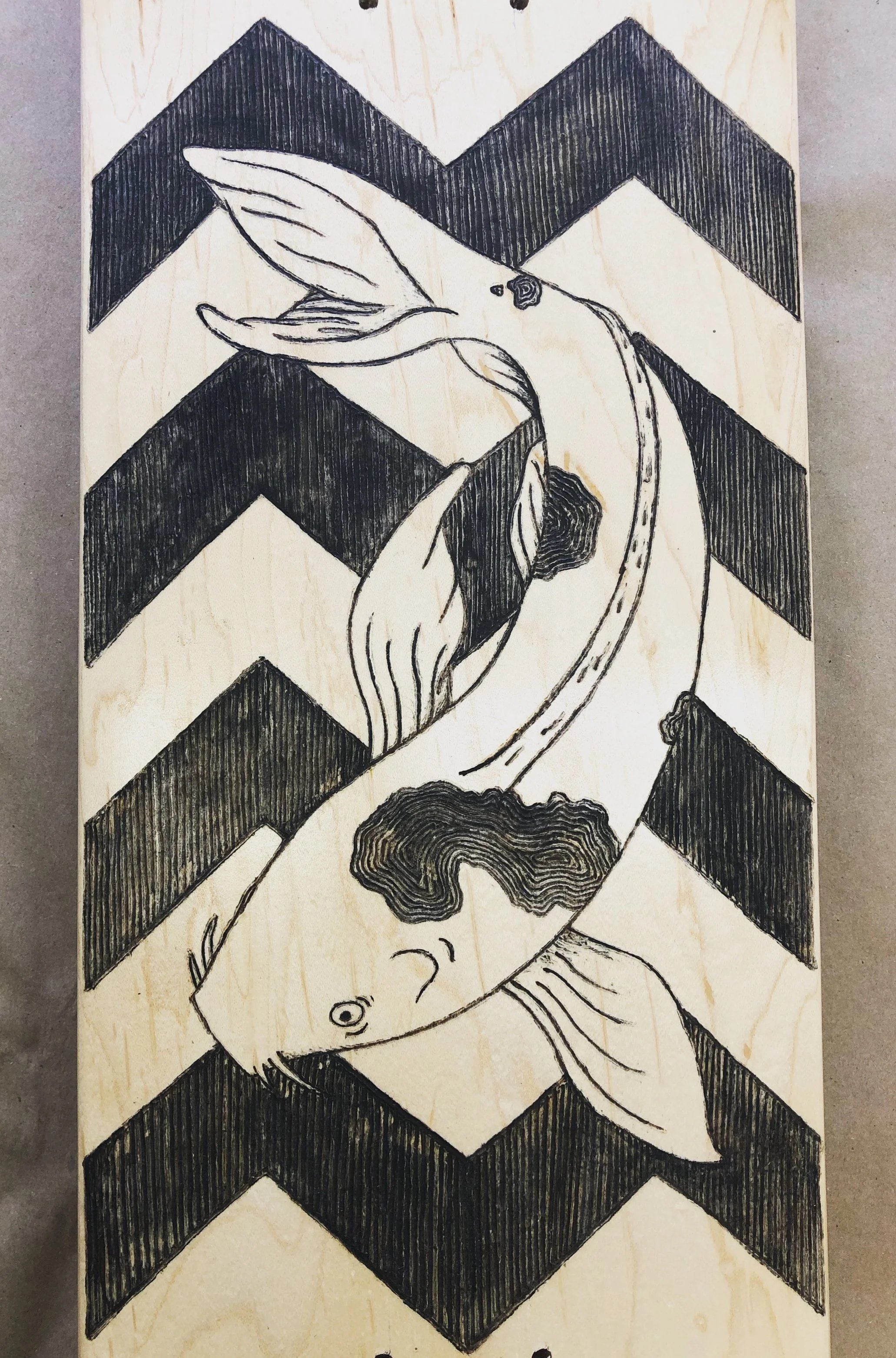 A wooden skateboard deck with a koi against a chevron-filled background wood burned into the surface