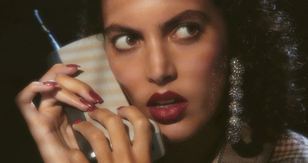 A woman with dark curly hair, wearing bold makeup including red lipstick and glittering earrings, holding a flip phone close to her ear.