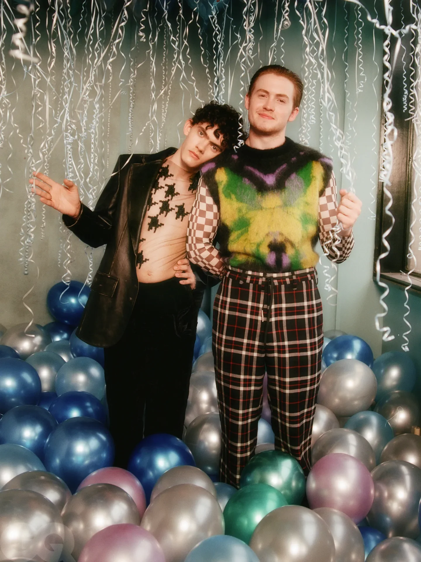 Two young men standing among colorful balloons and hanging silver streamers, celebrating at a party. One is wearing a leather jacket and mesh top with black cross patches, the other is in a colorful sweater and checkered pants.