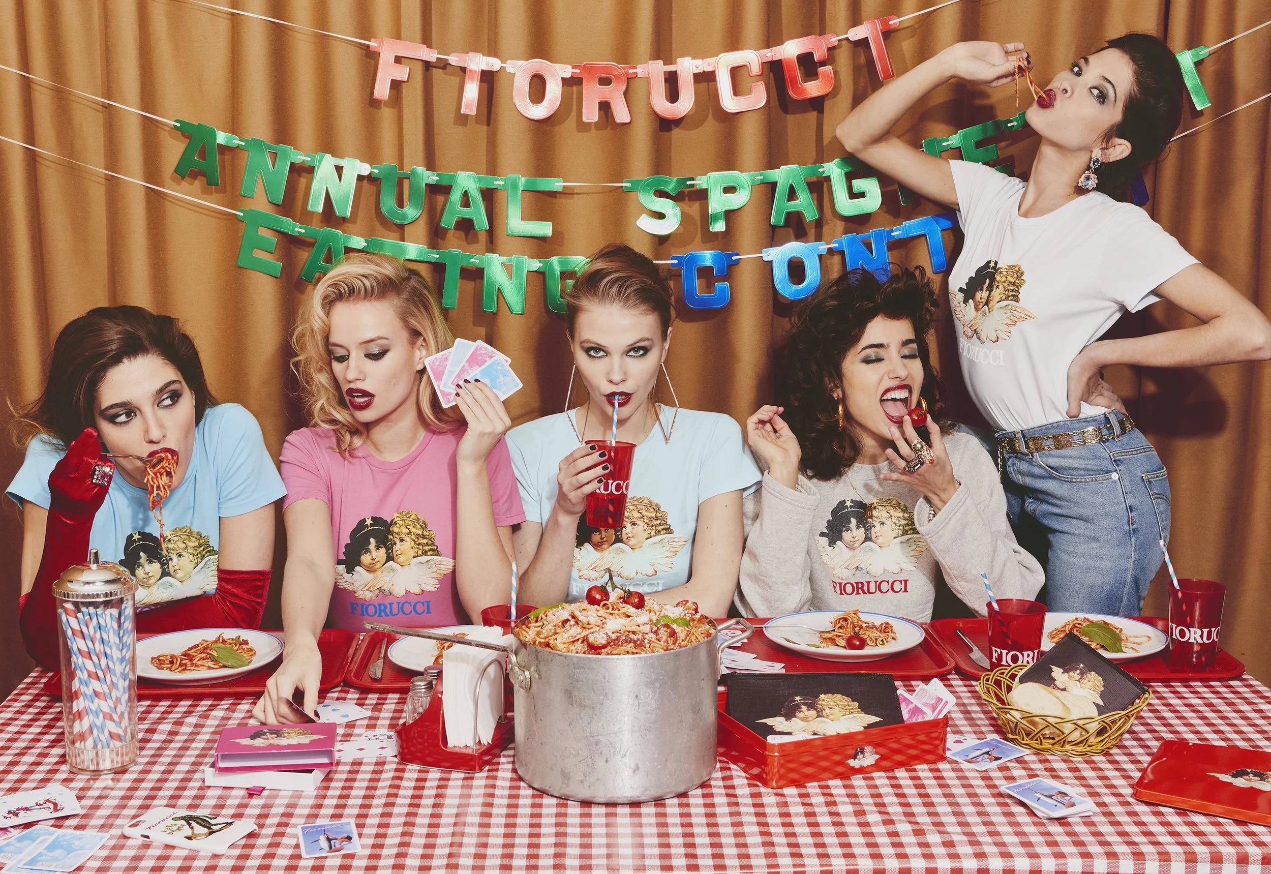 Five women sitting at a table with red and white checkered tablecloth, celebrating for Valentine's Day. They are wearing colorful T-shirts with an art design, and there are decorations in the background such as banners that read 'Fiorucci', 'Annual Spaghetti Eating Contest'. The table has plates of spaghetti, a large pot of spaghetti, cards, and drinks. One woman is drinking from a red cup, another is holding playing cards, and others are eating spaghetti.