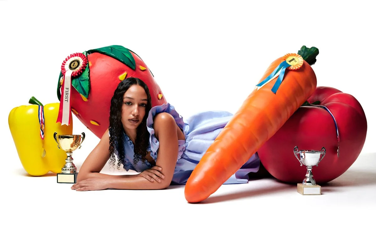 Young woman laying on a white surface surrounded by large colorful toy-like vegetables, including a yellow bell pepper, a strawberry, a large carrot, and a tomato, with some of them having ribbons and trophies.