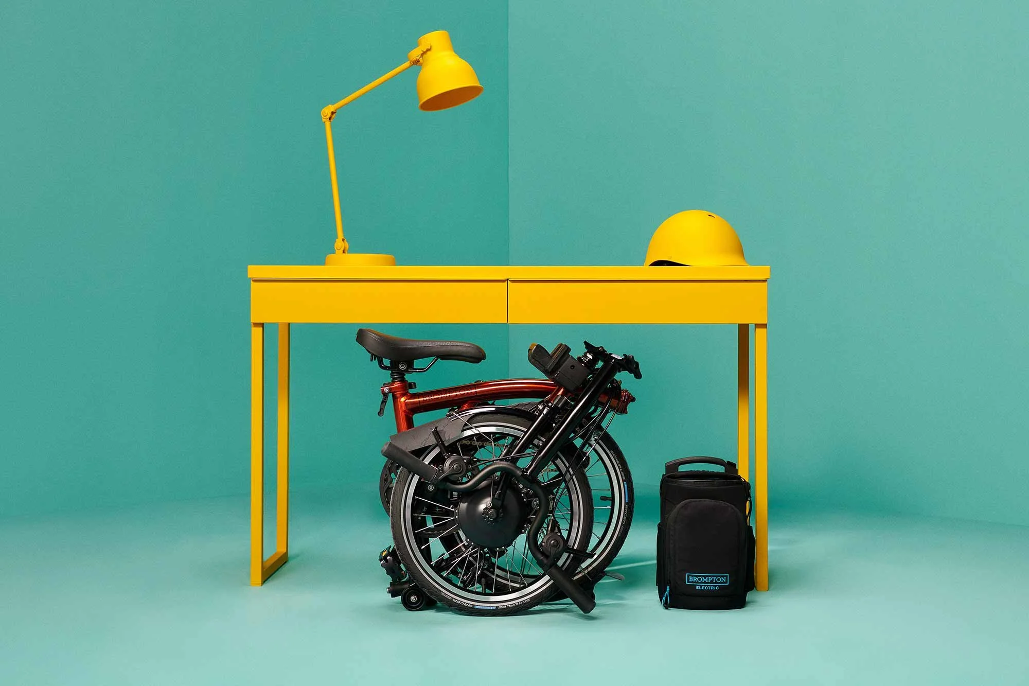 A yellow desk with a yellow desk lamp and a yellow helmet on top, a folded black electric bicycle with a red frame underneath, and a black Brompton backpack on a light blue background.