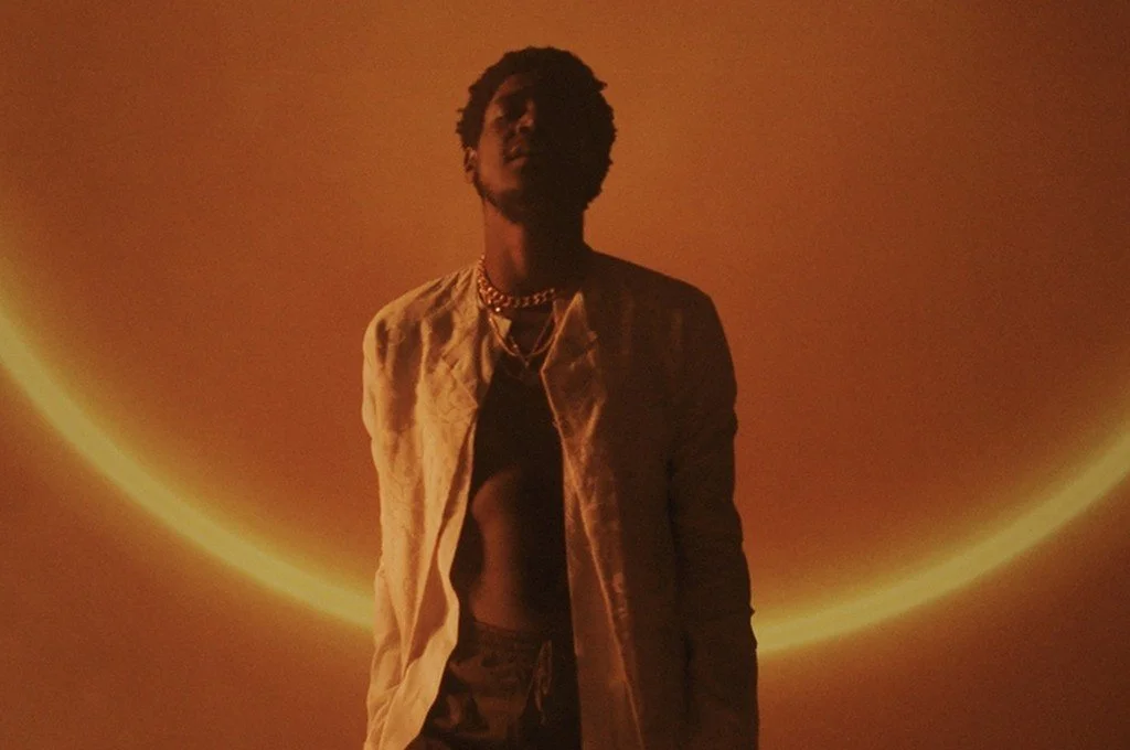 A person standing in front of a circular light on the wall, wearing a light-colored jacket and jewelry, with a dark background.