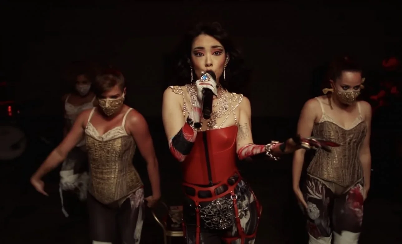 A woman in a red corset dress with jewelry, holding a microphone, is surrounded by four women wearing gold masks and corset tops in a dark setting.