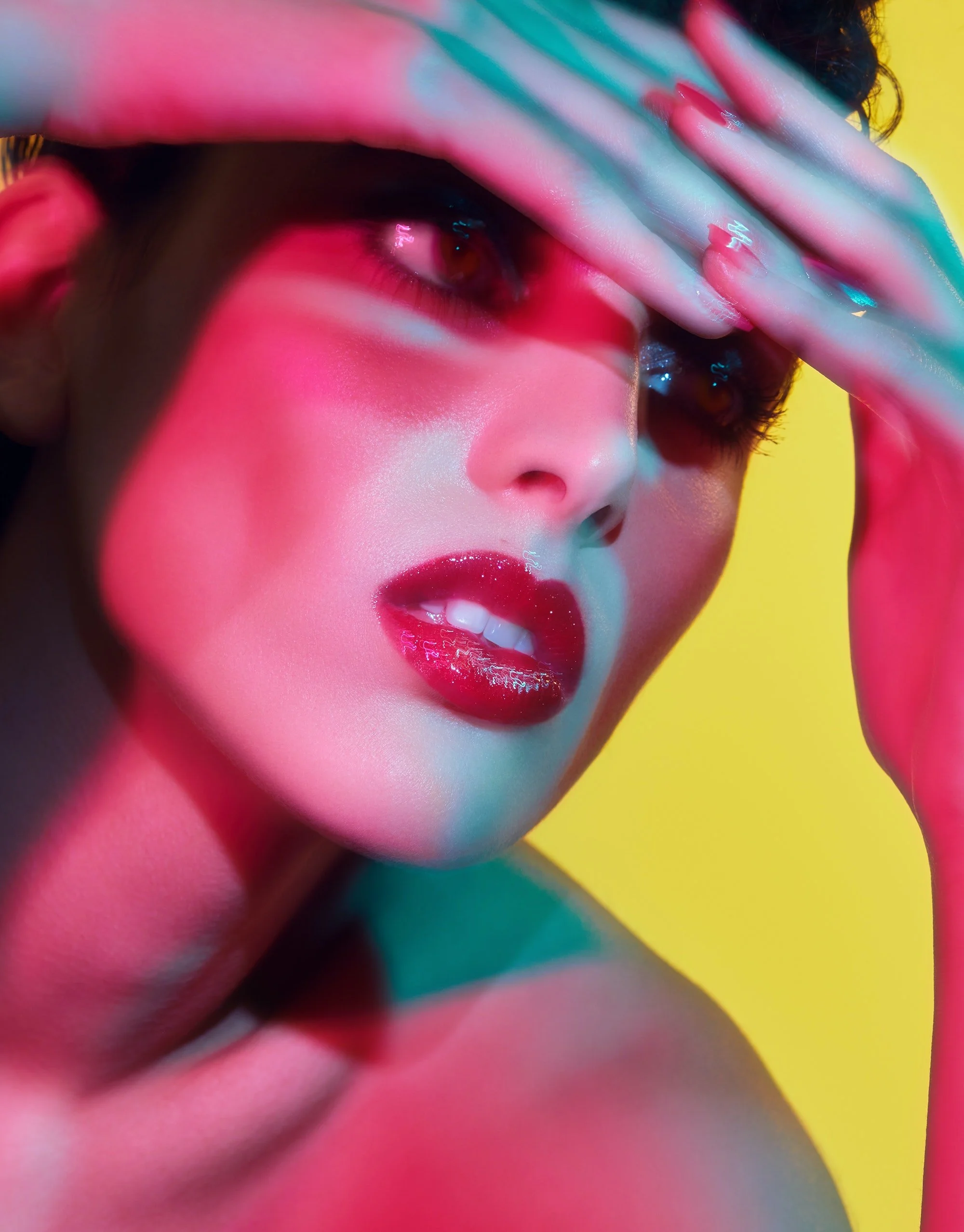 Close-up of a woman with bold makeup and colorful lighting, holding her hand over her forehead.