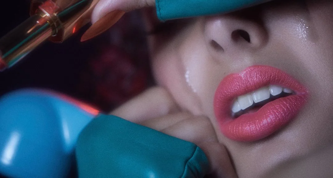 Close-up of a woman's face with pink lipstick, her lips slightly parted showing white teeth, as she lies on a surface with medical or cosmetic tools around her face.