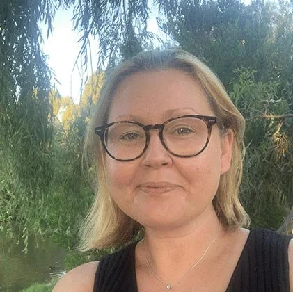 A woman with glasses and blonde hair taking a selfie outdoors near trees and a water body.