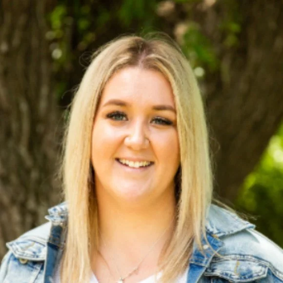 A smiling young woman with blonde hair wearing a denim jacket outdoors near a tree.