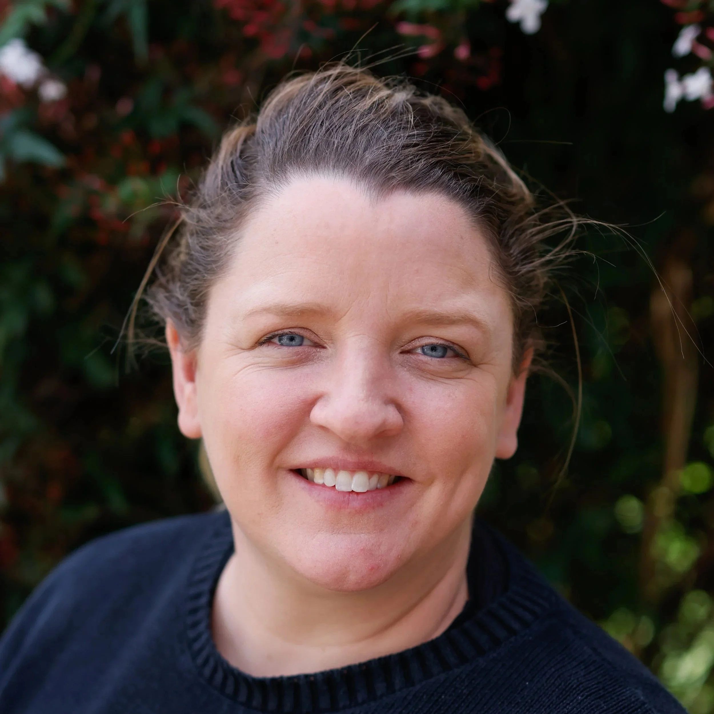 A woman smiling outdoors in front of green foliage, wearing a colorful patterned top.