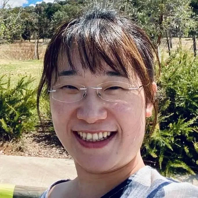 Smiling woman wearing glasses outdoors in a park on a sunny day, with trees and bushes in the background.