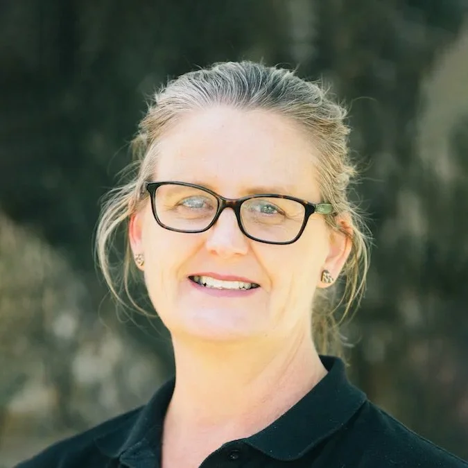 A woman with glasses, smiling, with light-colored hair tied back, wearing a black top, outdoors with blurred trees in the background.