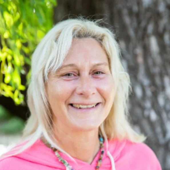 A smiling woman with blonde hair standing outdoors next to a tree, wearing a pink hoodie and a beaded necklace.