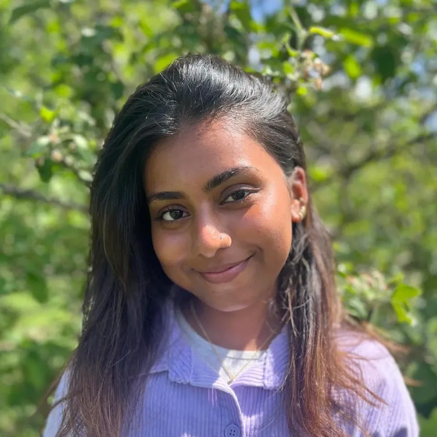 A young woman with long brown hair and a nose piercing, smiling outdoors with green foliage in the background.