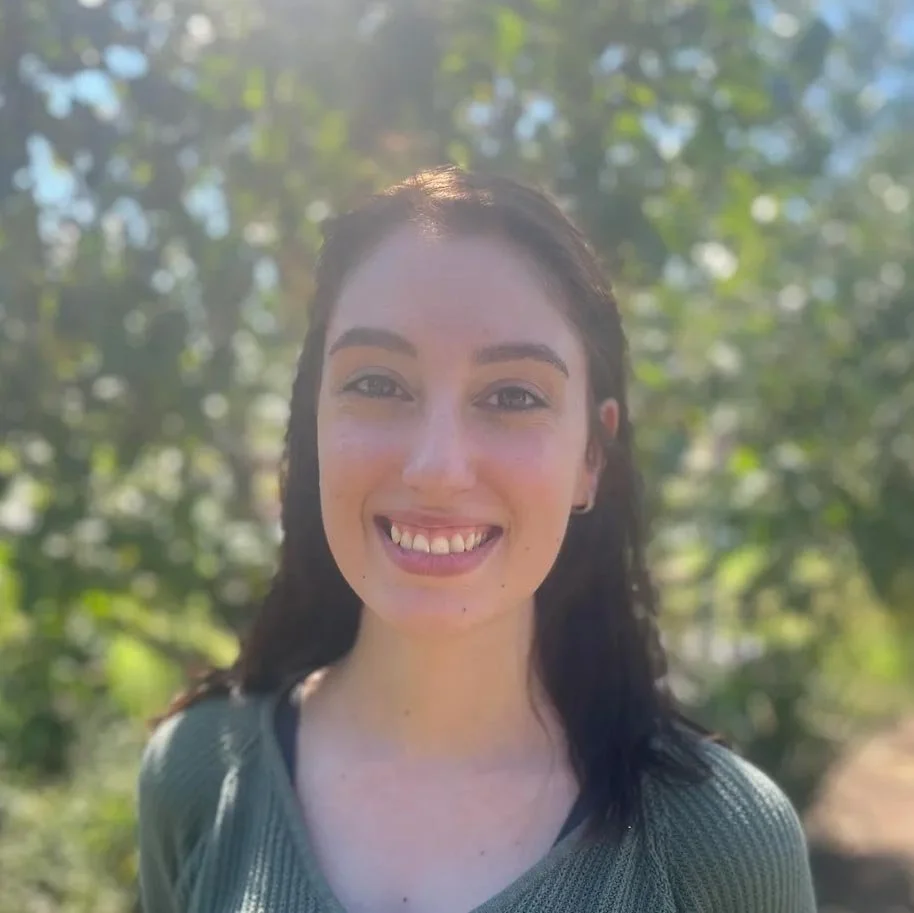 A woman smiling outdoors with sunlight and trees in the background.