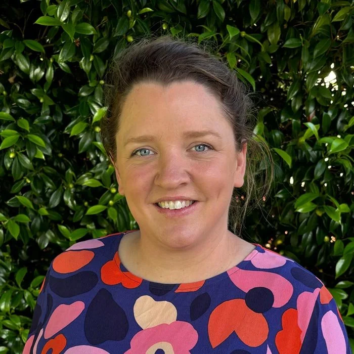 A woman smiling outdoors in front of green foliage, wearing a colorful patterned top.