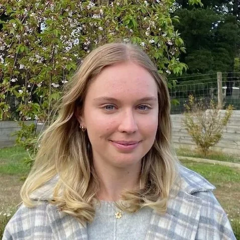Young woman with blonde hair and blue eyes smiling outdoors in front of flowering tree and fence.