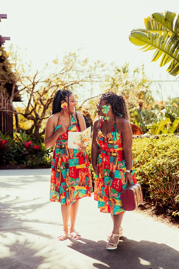 Two women in colorful tropical dresses walking and smiling outdoors, with one holding a map and the other carrying a vintage suitcase, surrounded by lush greenery and bright sunlight.