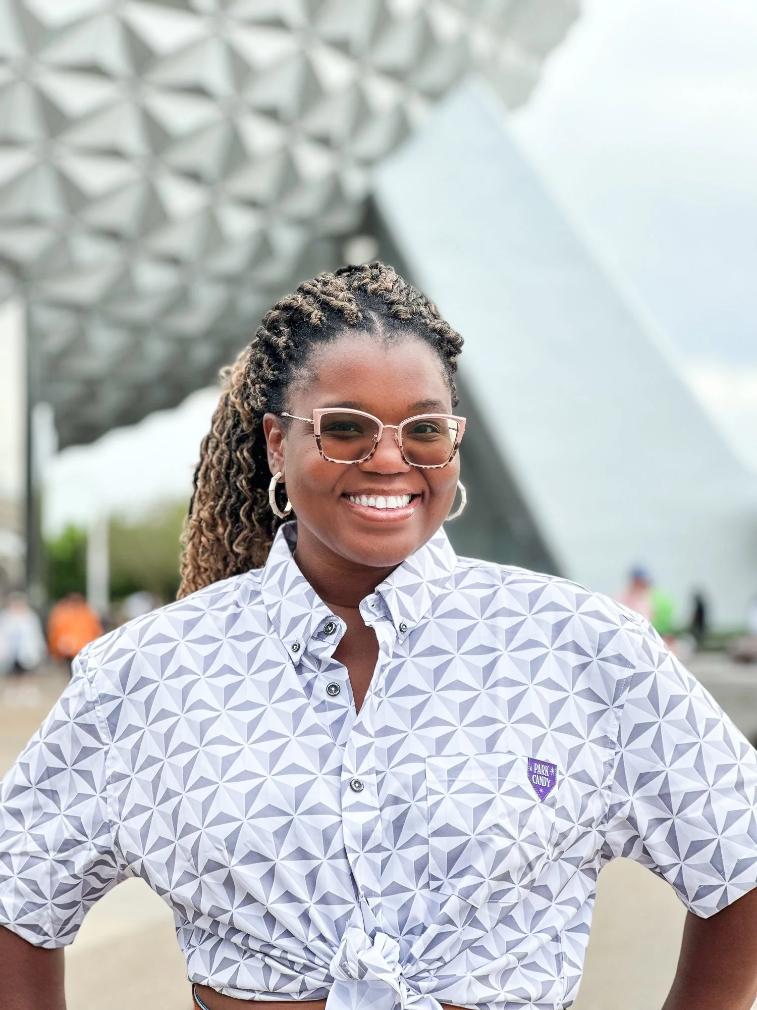 A smiling woman with braids, wearing sunglasses and hoop earrings, standing outdoors with the Space Needle in the background on a cloudy day.