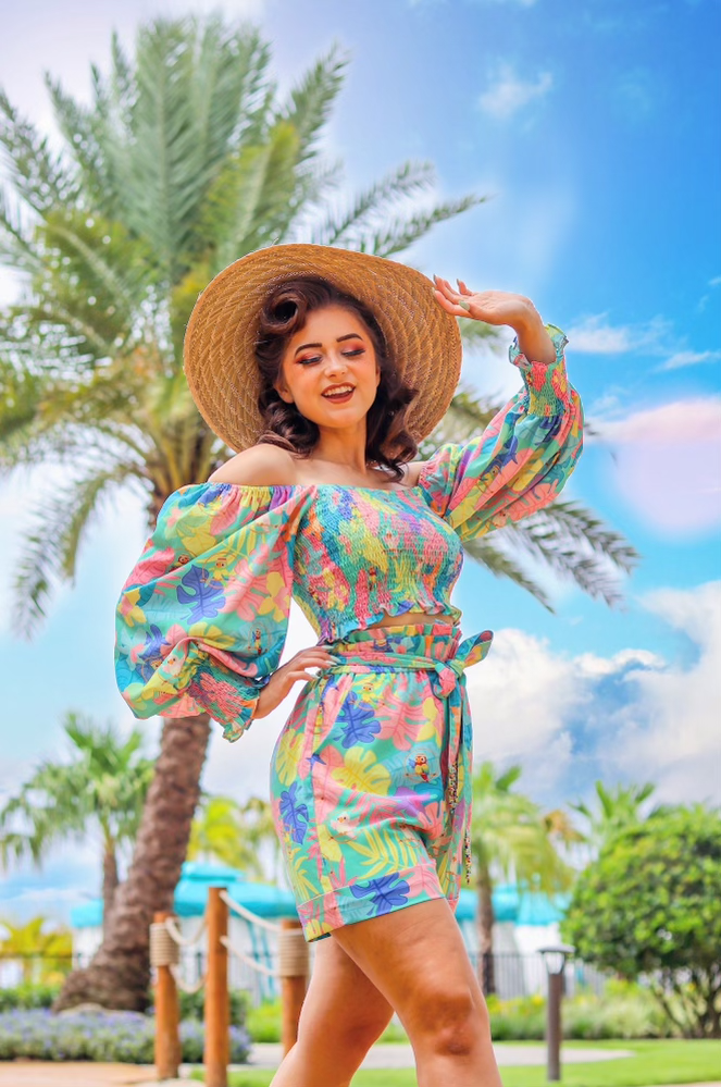 Woman wearing a colorful tropical dress and a large straw hat, standing outdoors with palm trees and a blue sky in the background.