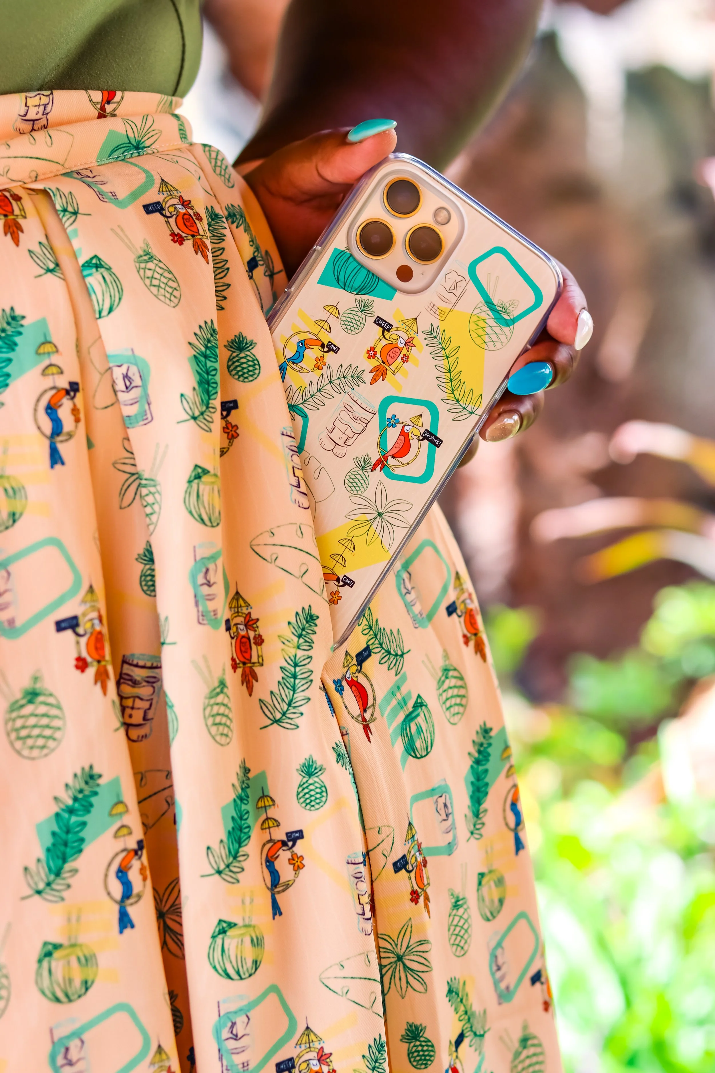 A person holding a clear phone case with tropical-themed stickers, wearing a peach-colored skirt with a pineapple, toucan, and flamingo pattern, outdoors with blurred greenery in the background.