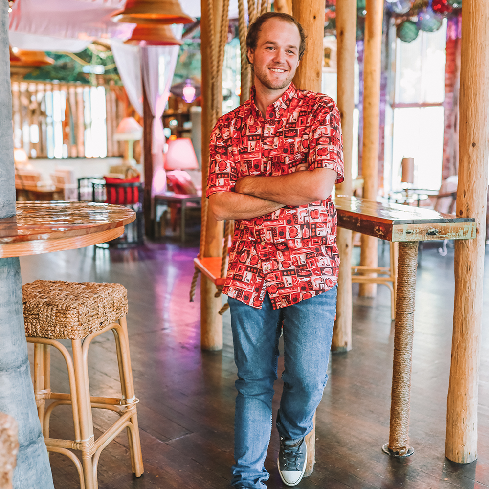 A young man with a slight beard and long hair tied back, smiling with arms crossed, standing inside a rustic restaurant or cafe with wooden beams and cozy decor.