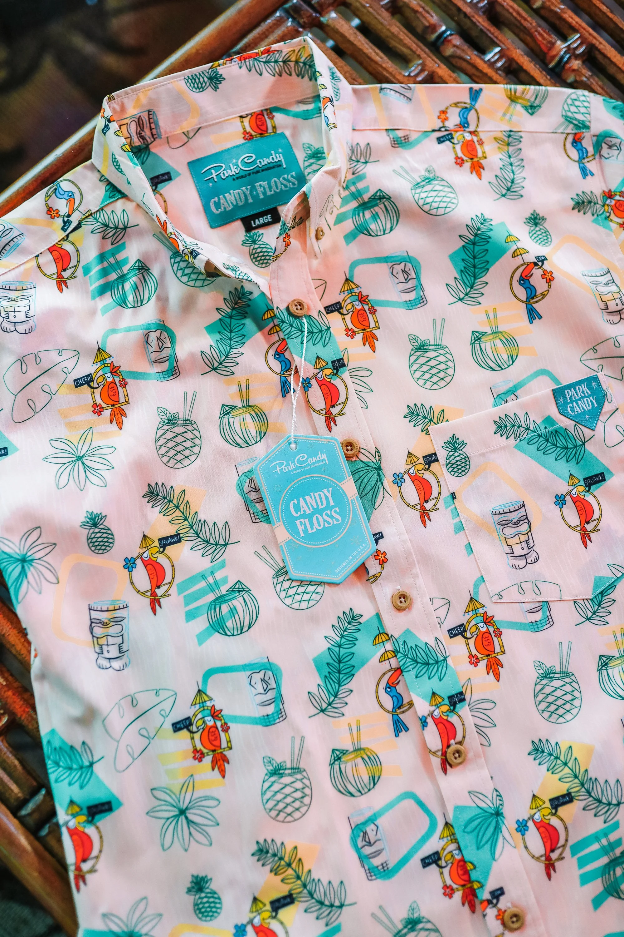 Colorful Hawaiian-style short-sleeve shirt with tropical prints of pineapples, leaves, and parrots, displayed on a wooden surface. The shirt has a tag that reads 'Candy Floss' and a small pocket on the chest.