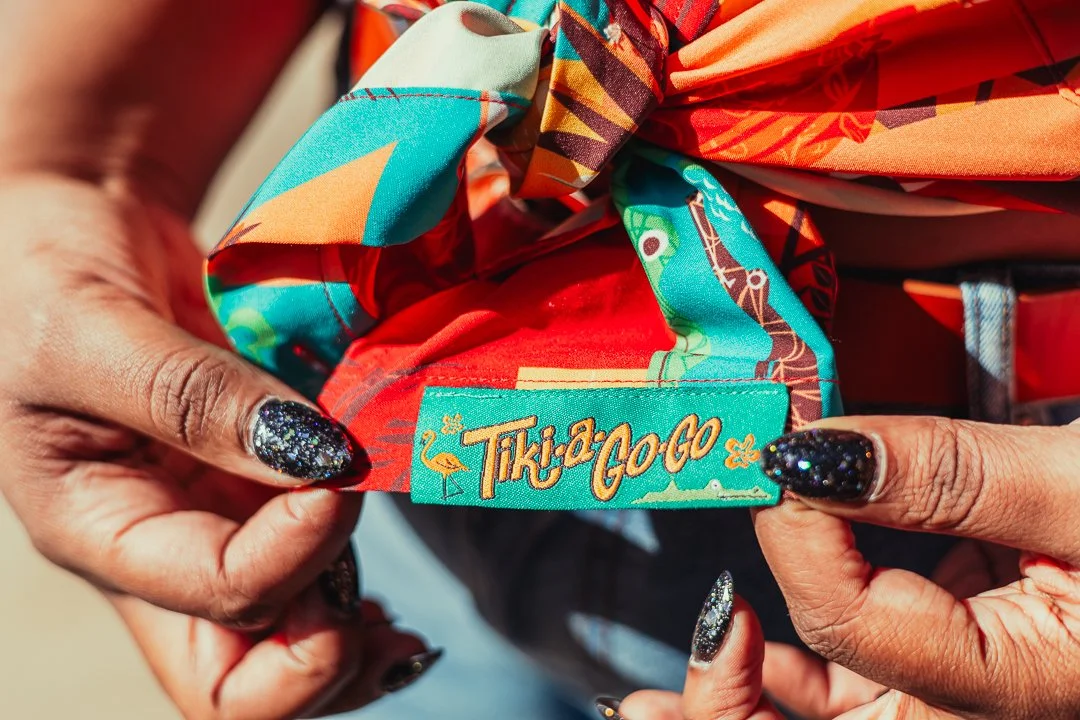 Close-up of someone's hands holding a colorful fabric with a "Tiki-Go-Go" label, featuring bright tropical-themed designs and patterns.