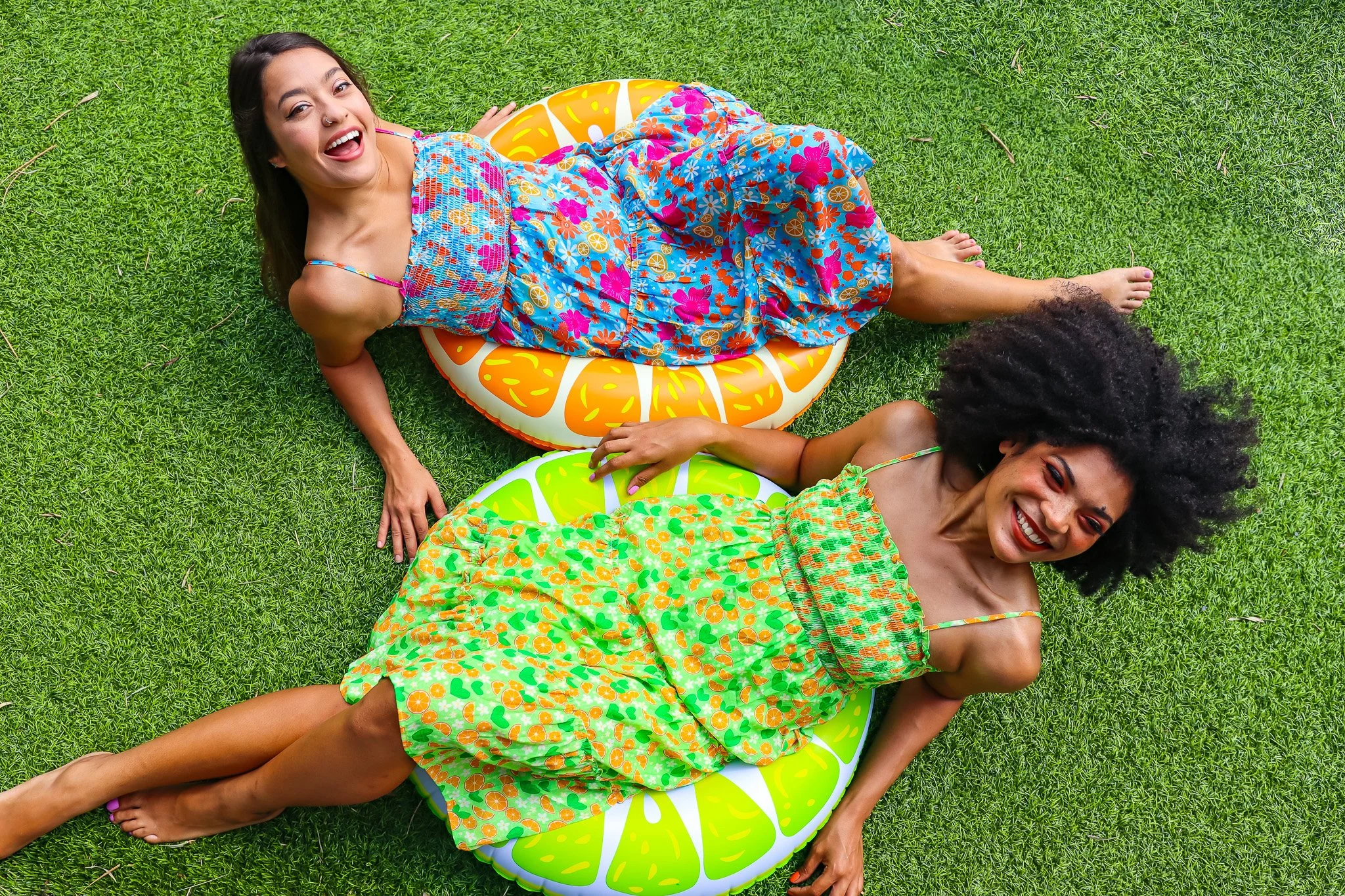 Two women in colorful dresses lying on inflatable pool floats shaped like citrus slices, on green grass, smiling and enjoying outdoor summer weather.
