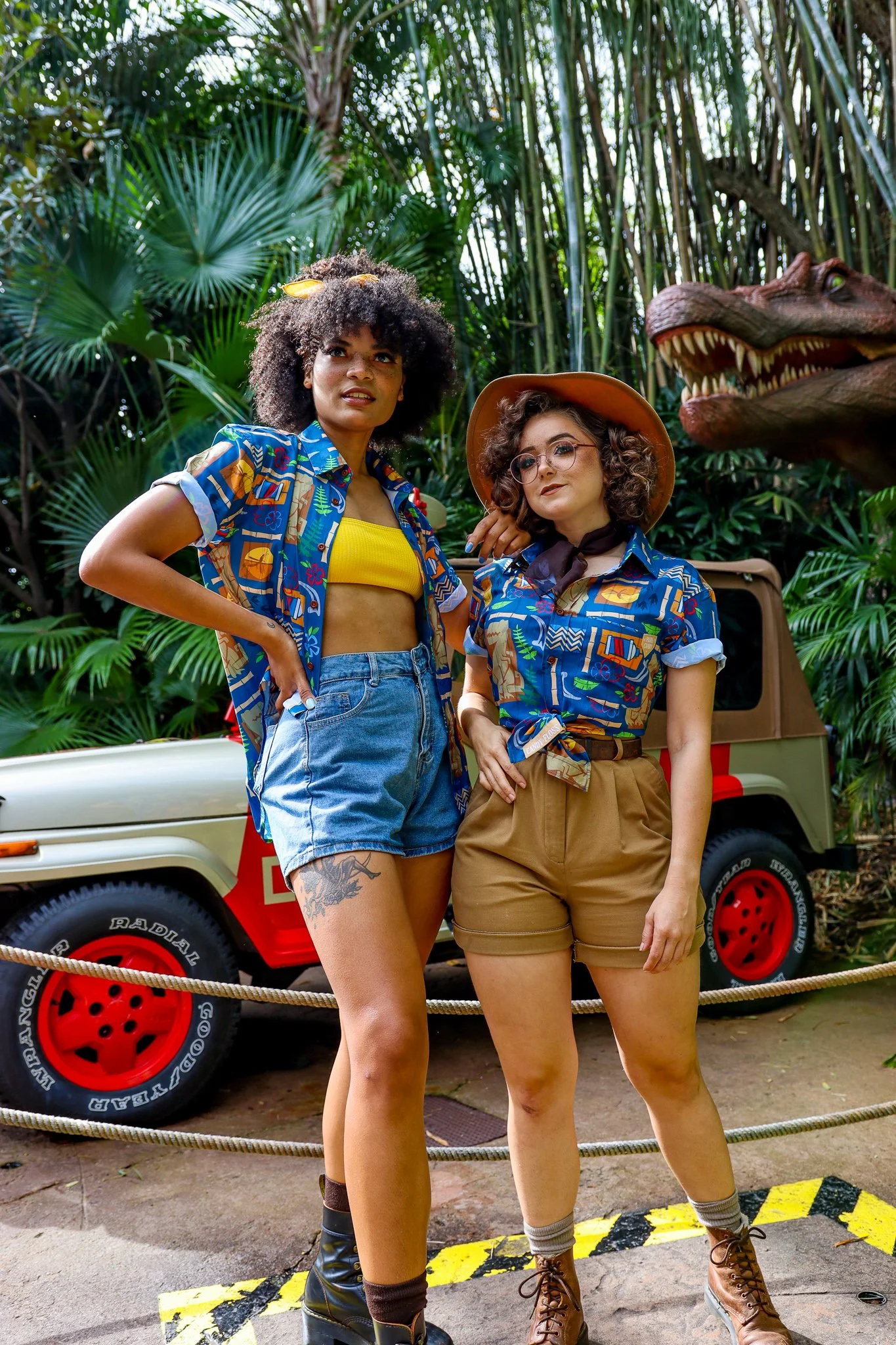 Two women dressed in jungle-themed outfits stand in front of a dinosaur figure and a jungle vehicle. One woman, with curly hair and darker skin, wears a colorful shirt, yellow crop top, denim shorts, and has tattoos on her thigh. The other woman, wit