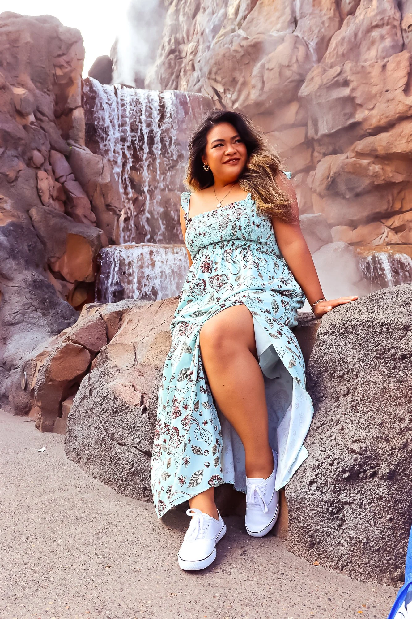 A woman sitting on a rock in front of a waterfall, wearing a blue dress with floral patterns and white sneakers, smiling and looking to the side.