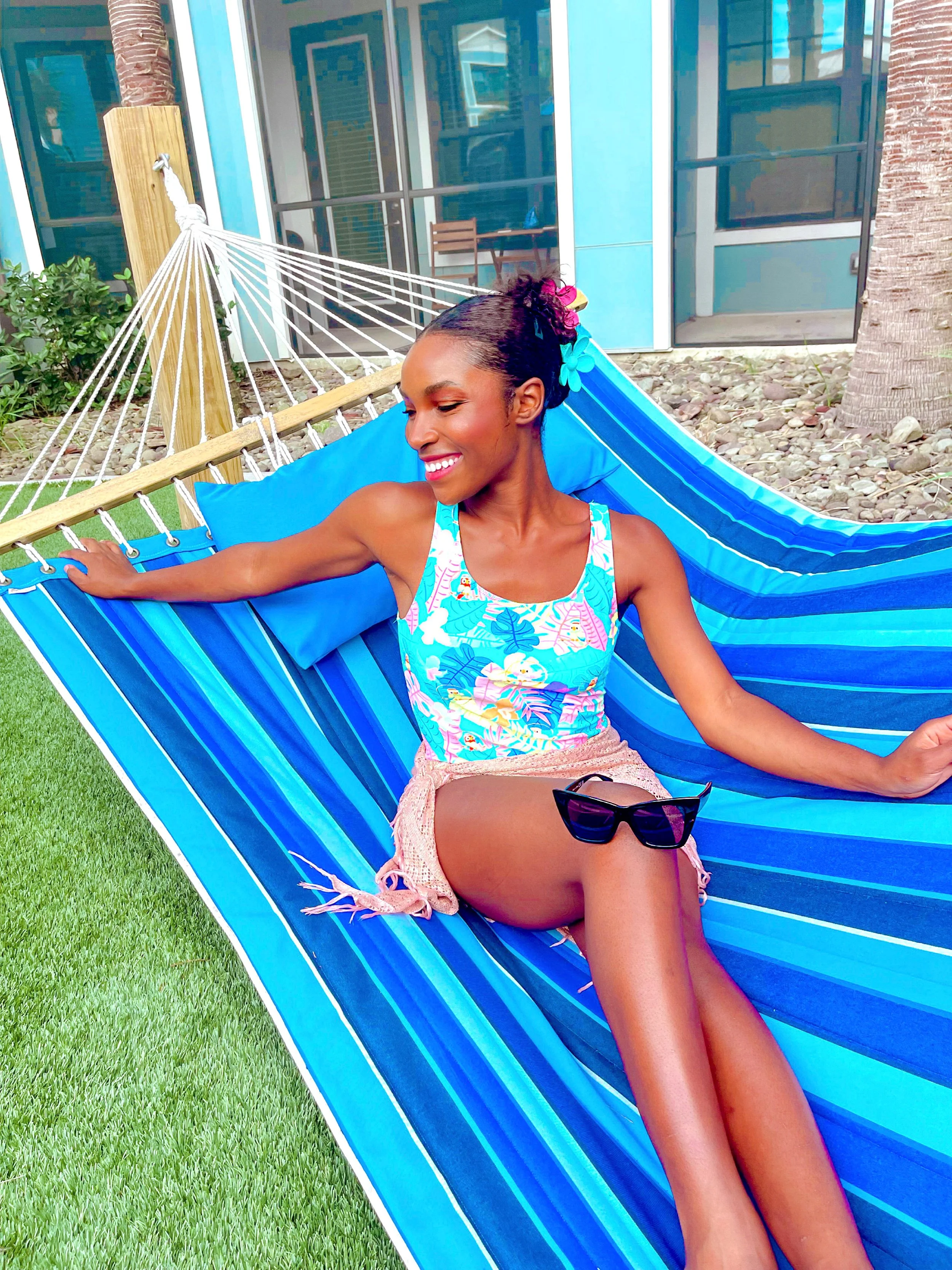 Smiling woman relaxing on a blue striped hammock outdoors near a building with glass doors, wearing a colorful tropical tank top, beige shorts, and sunglasses resting on her lap.