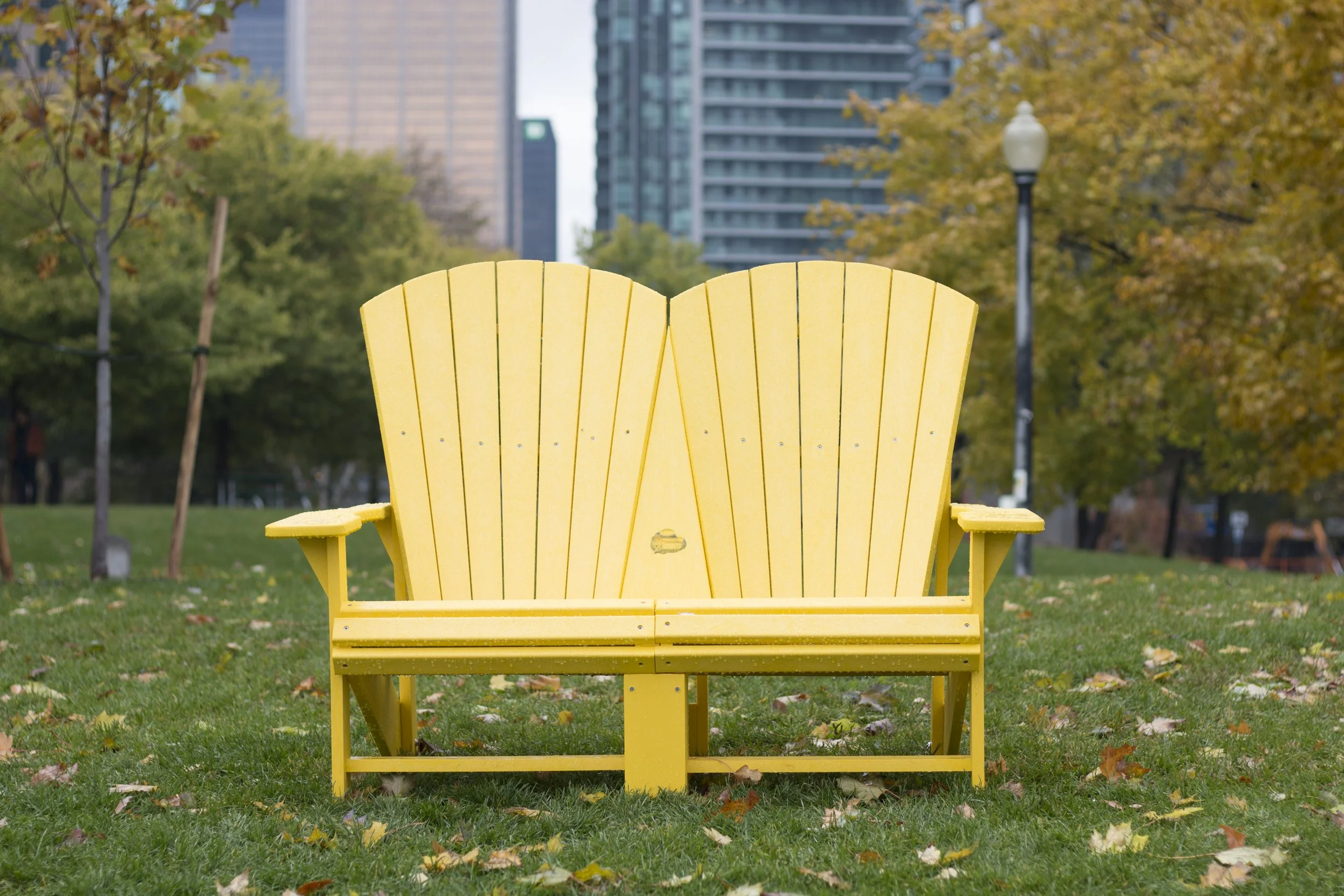 A bright yellow Adirondack style bench in a park with grass, trees with fall foliage, city buildings, and streetlamp in the background.