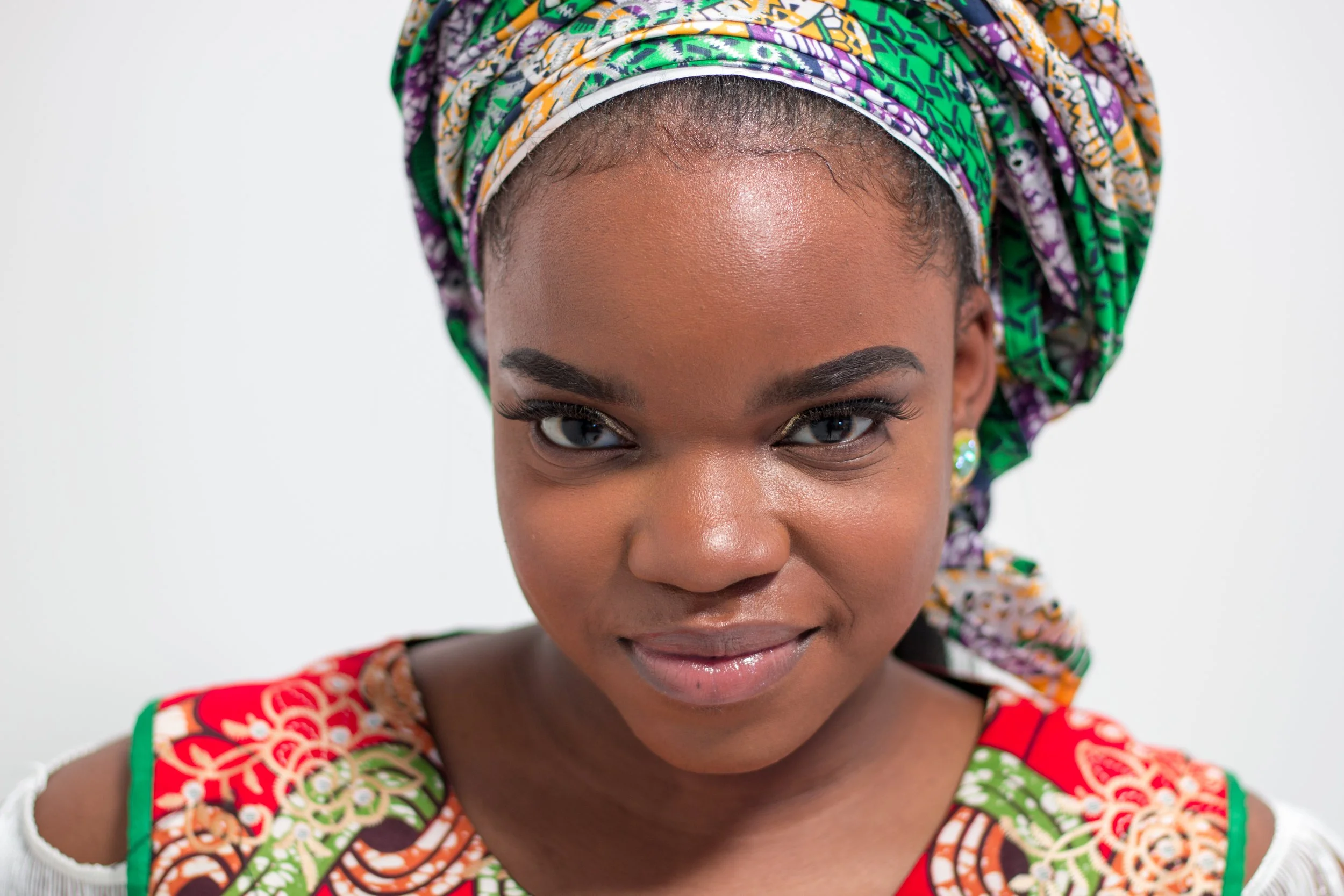 A close-up portrait of an African woman wearing a colorful, patterned headwrap and traditional dress, smiling gently with her shoulders exposed.