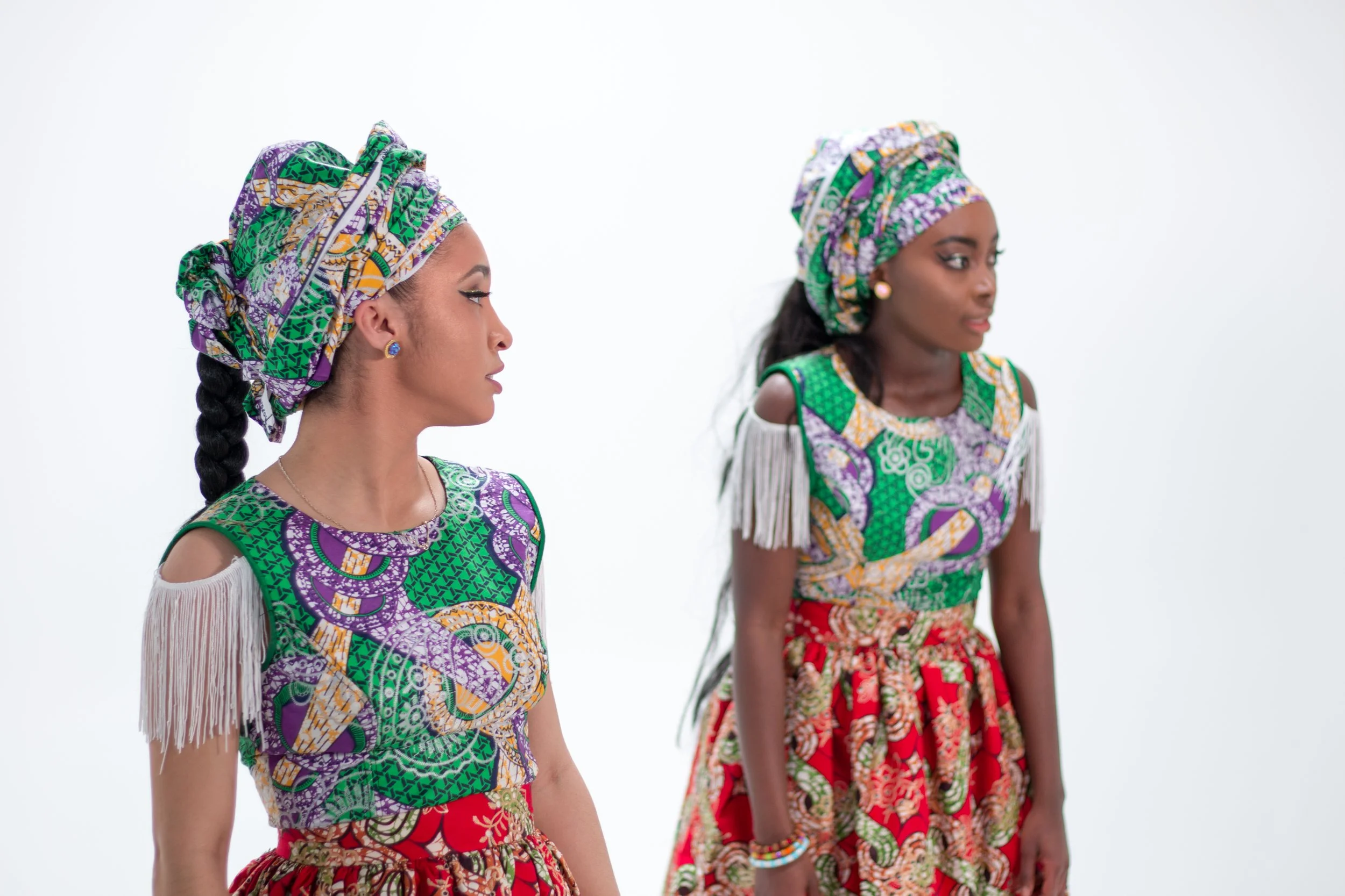 Two women dressed in colorful traditional African attire with head wraps, standing against a plain white background.