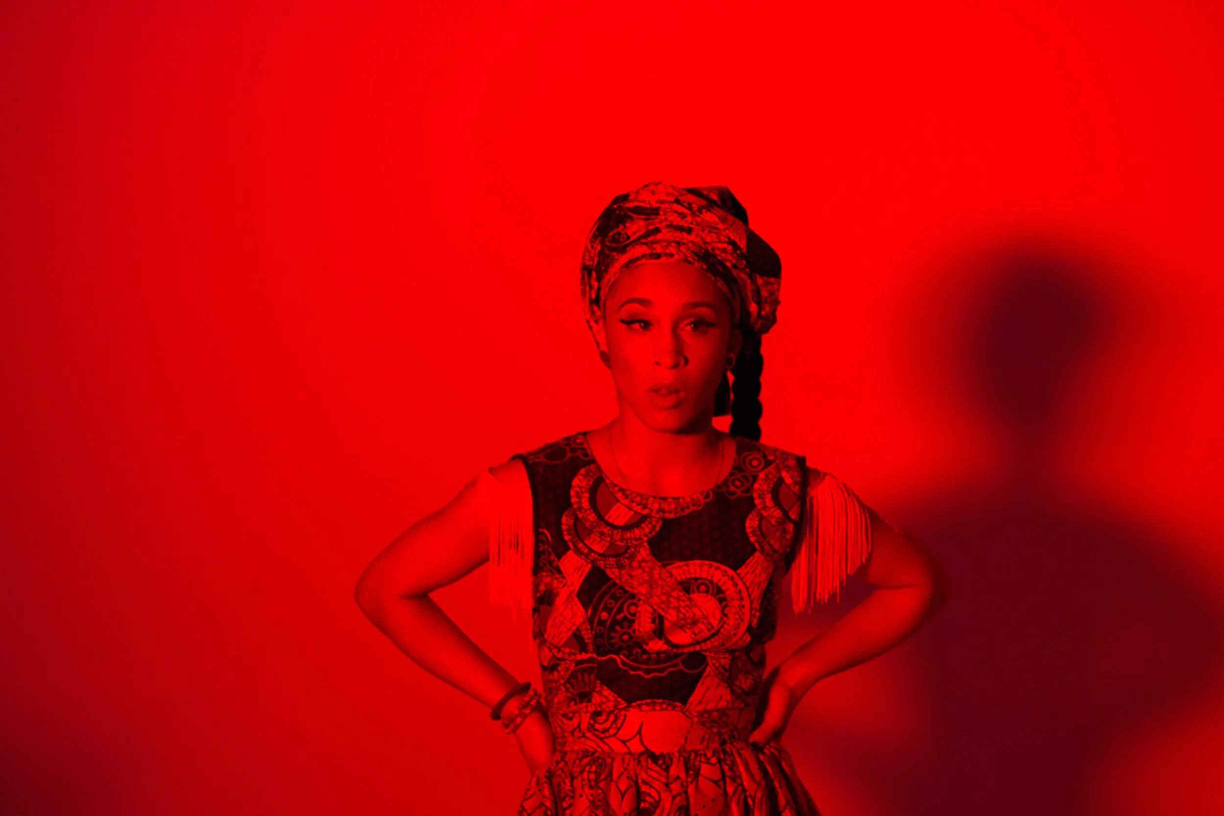 A woman wearing a patterned dress and headwrap stands against a red background, with her hands on her hips and an expressive look on her face.
