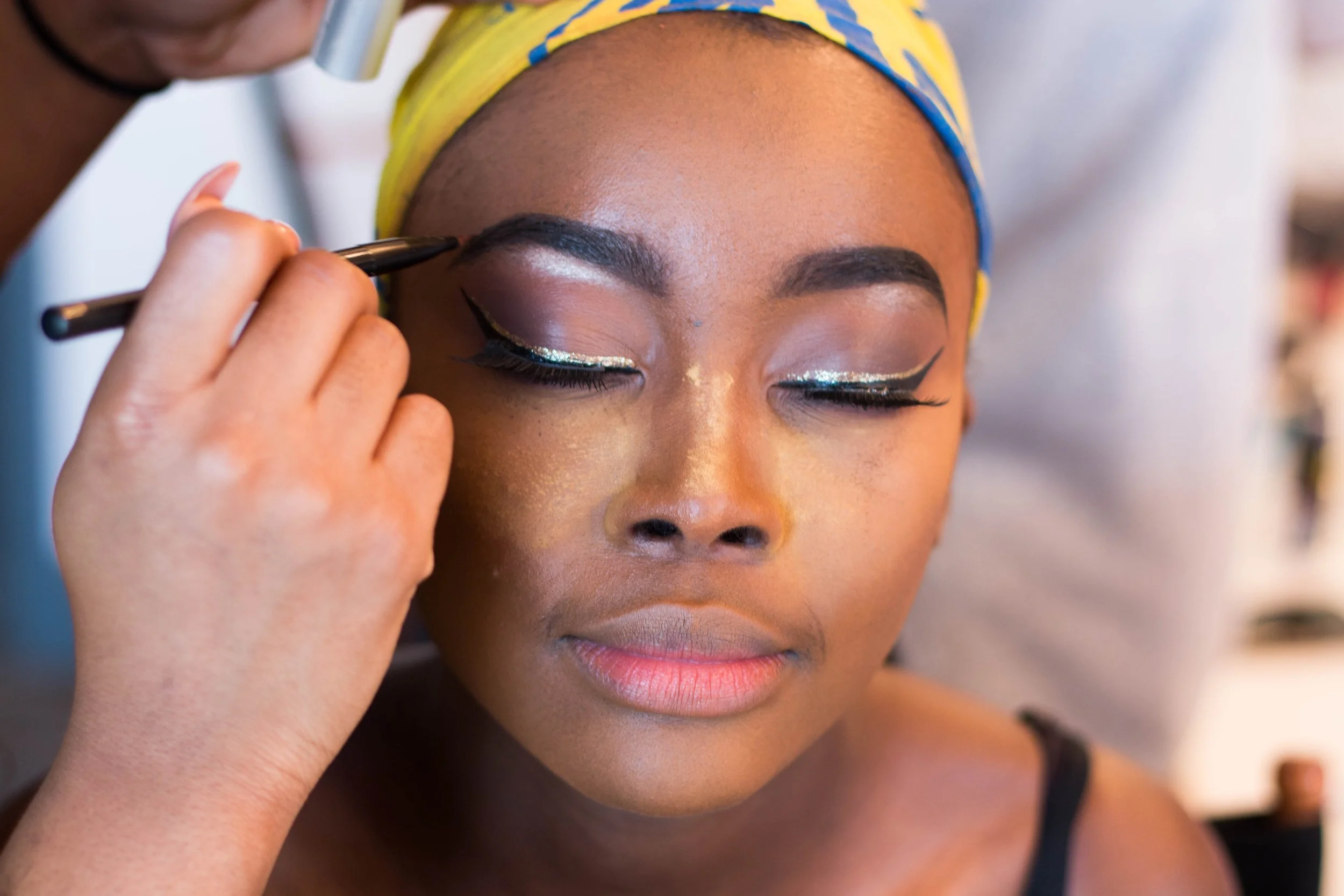 A woman with a colorful headscarf getting her makeup done, focusing on eye makeup with silver and black eyeliner.